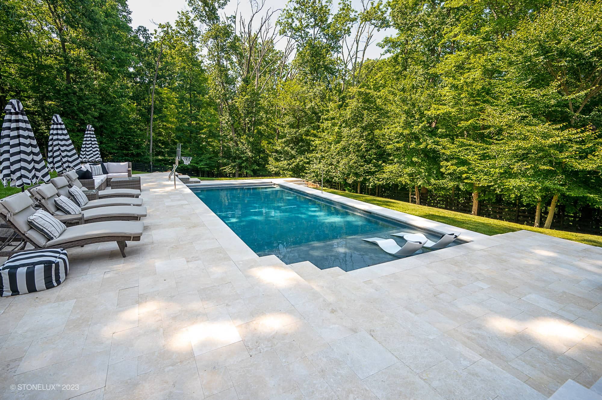 A rectangular outdoor swimming pool with clear water is surrounded by Pera Cream Marble tiles, lounge chairs, striped umbrellas, and greenery from tall trees in the background. Two pool loungers sit partially in the shallow end among the trees.