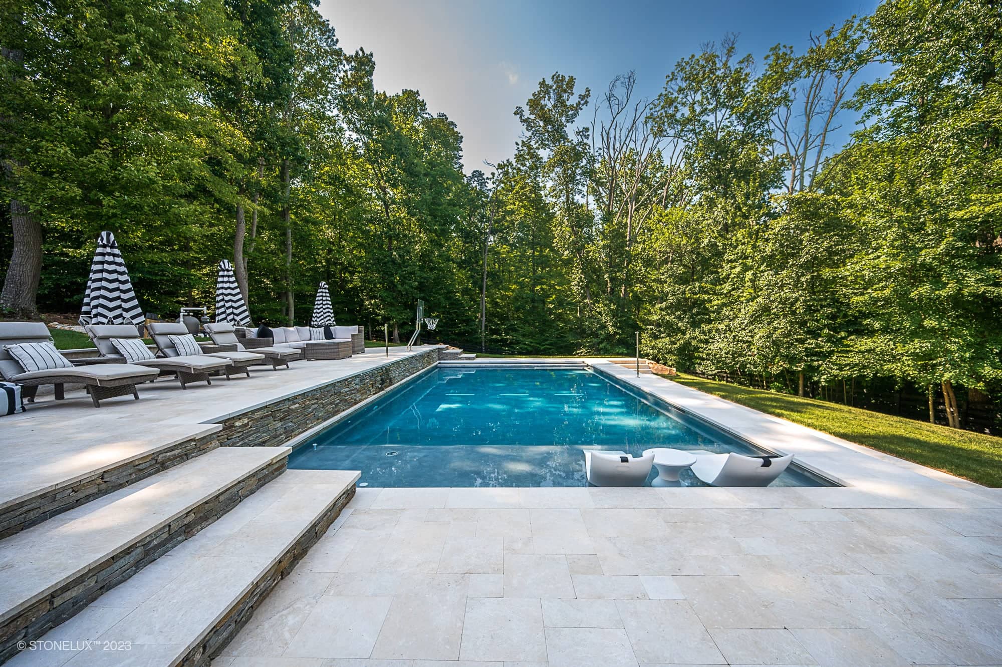 A luxurious outdoor pool surrounded by Pera Cream Marble tiles, lounge chairs, and black-and-white striped umbrellas, set among the trees of a lush green forest under a clear blue sky.