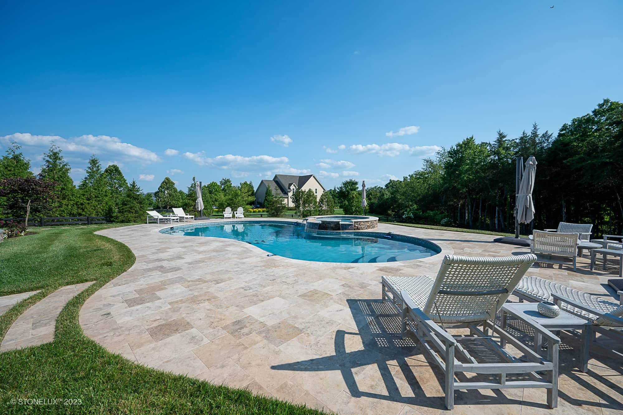 A luxurious backyard features a Travertine Charm: Philly Travertine Pool with beige and blue accents, a hot tub, lounge chairs, and umbrellas on a tiled patio, all surrounded by green lawn, trees, and a large house under a clear blue sky.