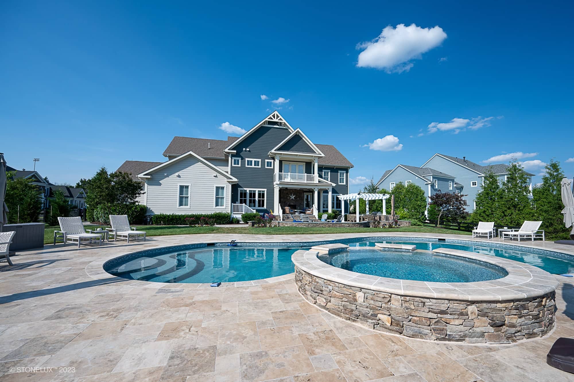 A large, modern house with a landscaped backyard featuring a Travertine Charm: Philly Travertine Pool with beige and blue accents, a round stone hot tub, lounge chairs, and lush greenery under a clear blue sky.
