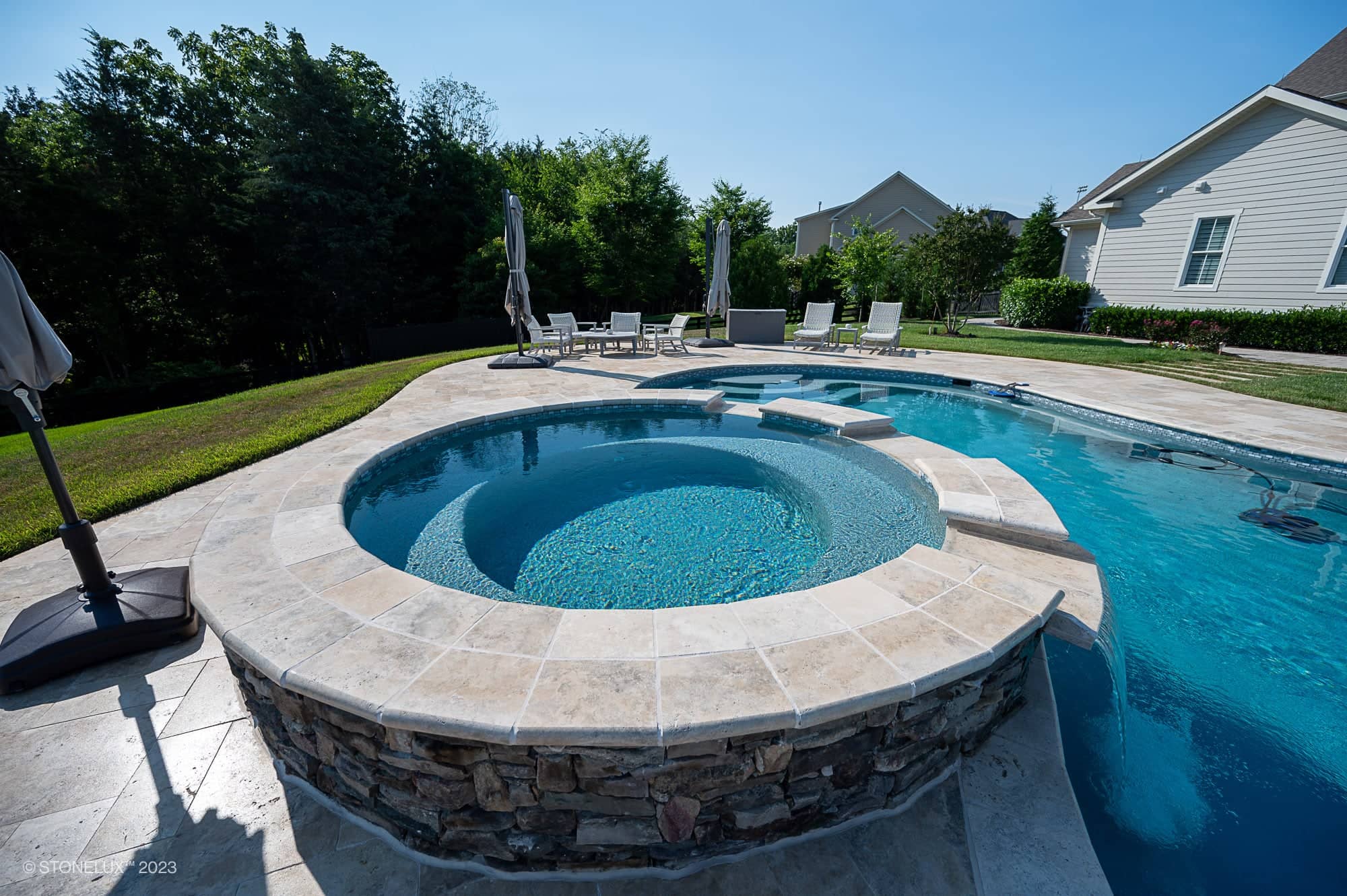 A backyard pool area with Travertine Charm: Philly Travertine Pool featuring beige and blue accents, a circular hot tub connected to the pool, surrounded by stone tiles, lounge chairs, umbrellas, lush grass and trees.