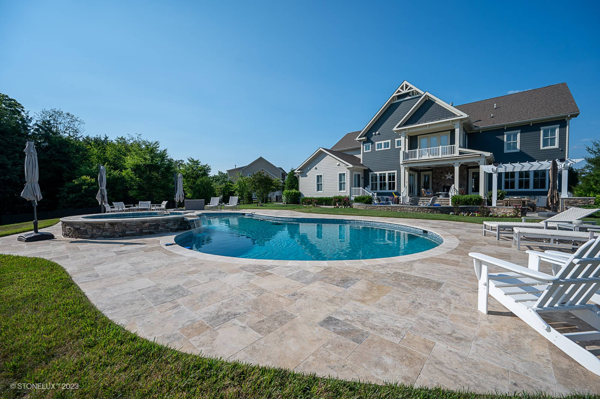 A modern house with a large backyard featuring a Travertine Charm: Philly Travertine Pool with beige and blue accents, a hot tub, patio seating, lounge chairs, and green lawn under a clear blue sky.