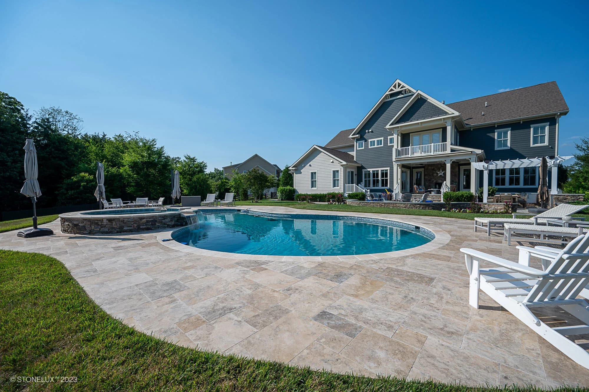 A modern house with a large backyard featuring a Travertine Charm: Philly Travertine Pool with beige and blue accents, a hot tub, patio seating, lounge chairs, and green lawn under a clear blue sky.