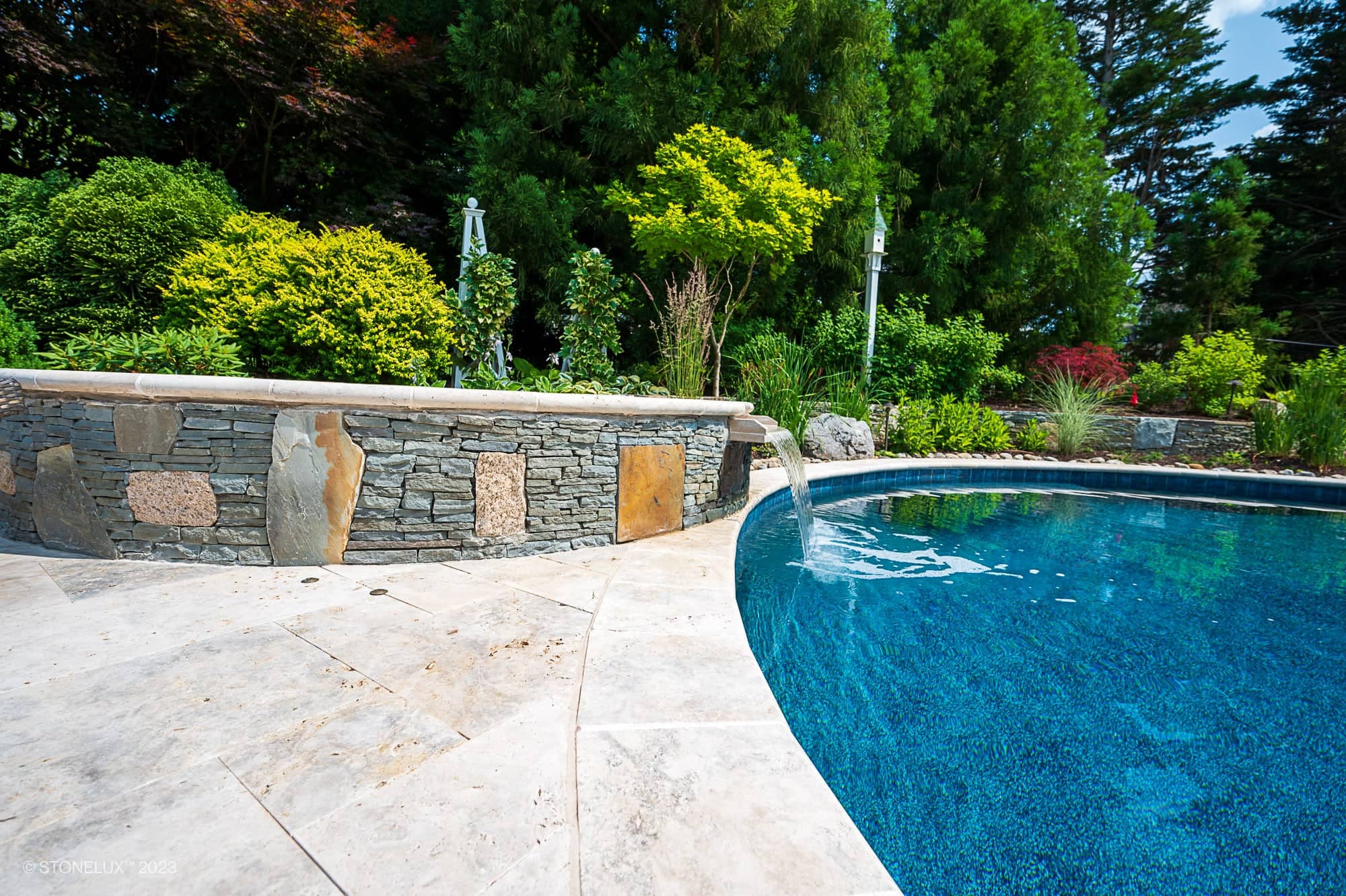 A curved stone wall in Travertine Serenity shades of beige, silver, and brown features a small water spout flowing into a clear blue swimming pool, surrounded by lush green trees and bushes on a sunny day.