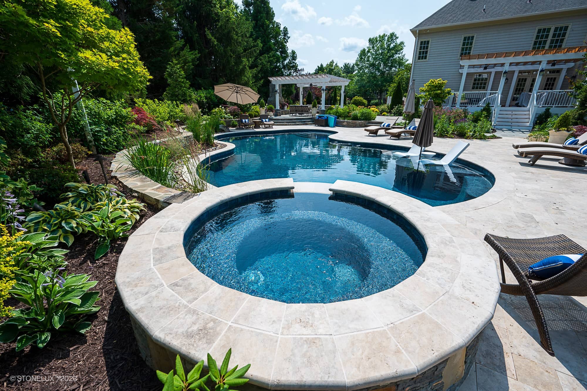 A luxurious backyard features a round brown and silver hot tub connected to a large, curvy swimming pool framed with travertine serenity—a poolside palette of beige stone, lounge chairs, plants, and a pergola beside the house on a sunny day.