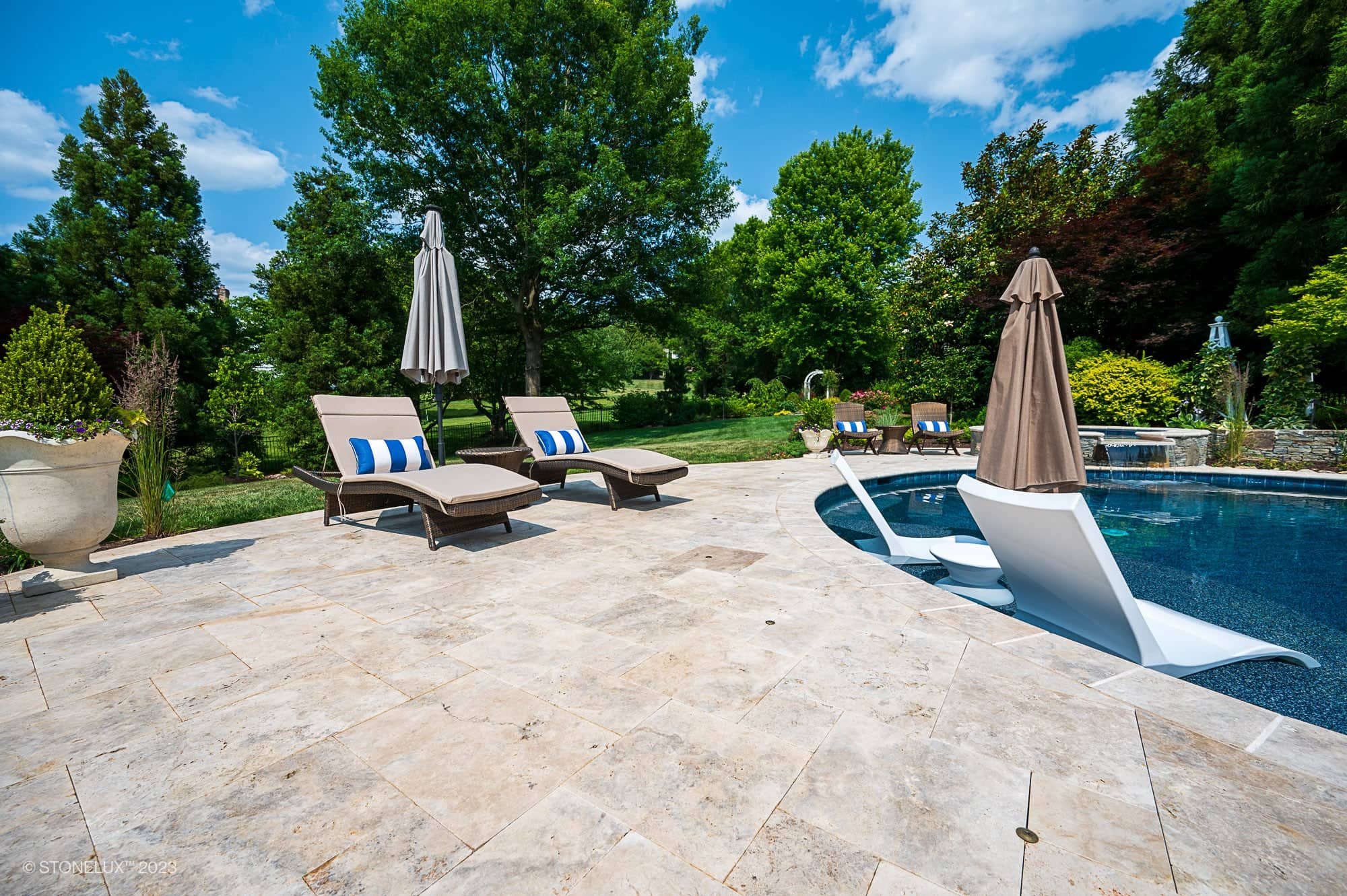 A sunny backyard with brown and silver accents features two lounge chairs and umbrellas by a stone-tiled pool. Travertine Serenity: A Poolside Palette of Beige is showcased as white loungers sit partially submerged, surrounded by lush greenery.