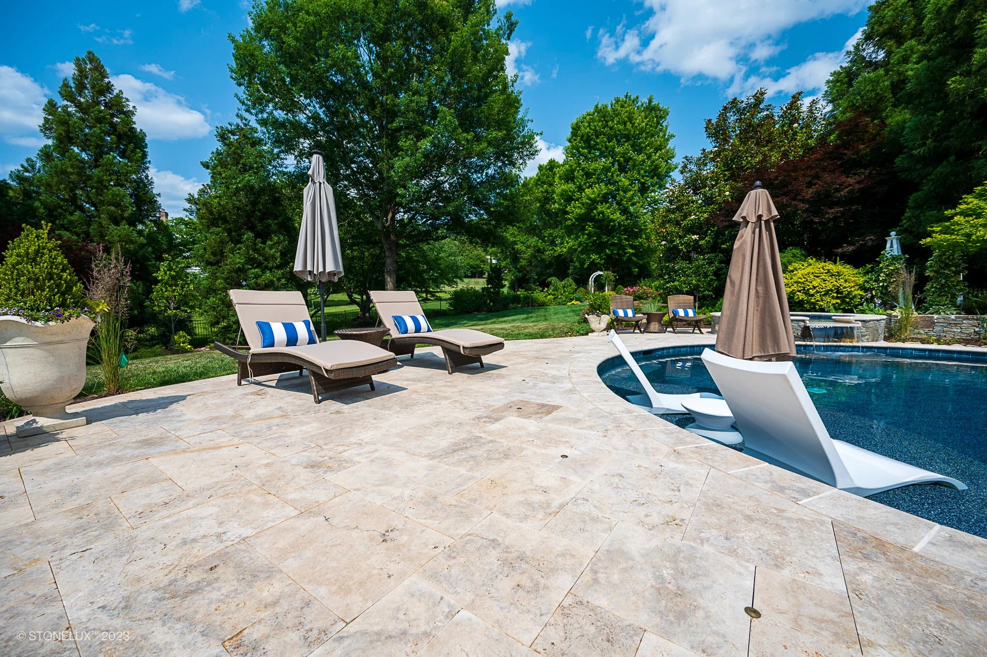 A sunny backyard with brown and silver accents features two lounge chairs and umbrellas by a stone-tiled pool. Travertine Serenity: A Poolside Palette of Beige is showcased as white loungers sit partially submerged, surrounded by lush greenery.