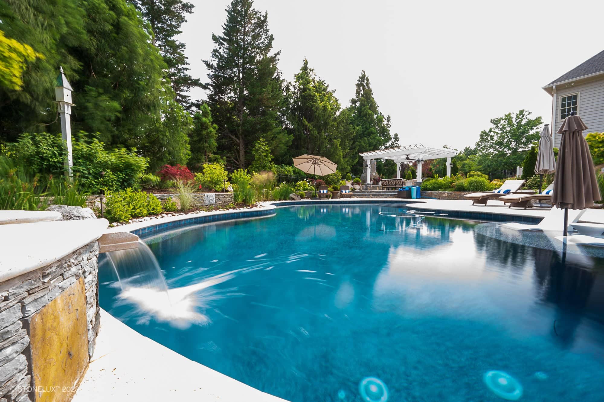 A luxurious backyard features a curved swimming pool with a small waterfall, surrounded by lounge chairs, umbrellas, and Silver accents atop travertine serenity—a poolside palette of beige, brown, lush greenery, and a white pergola in the background.
