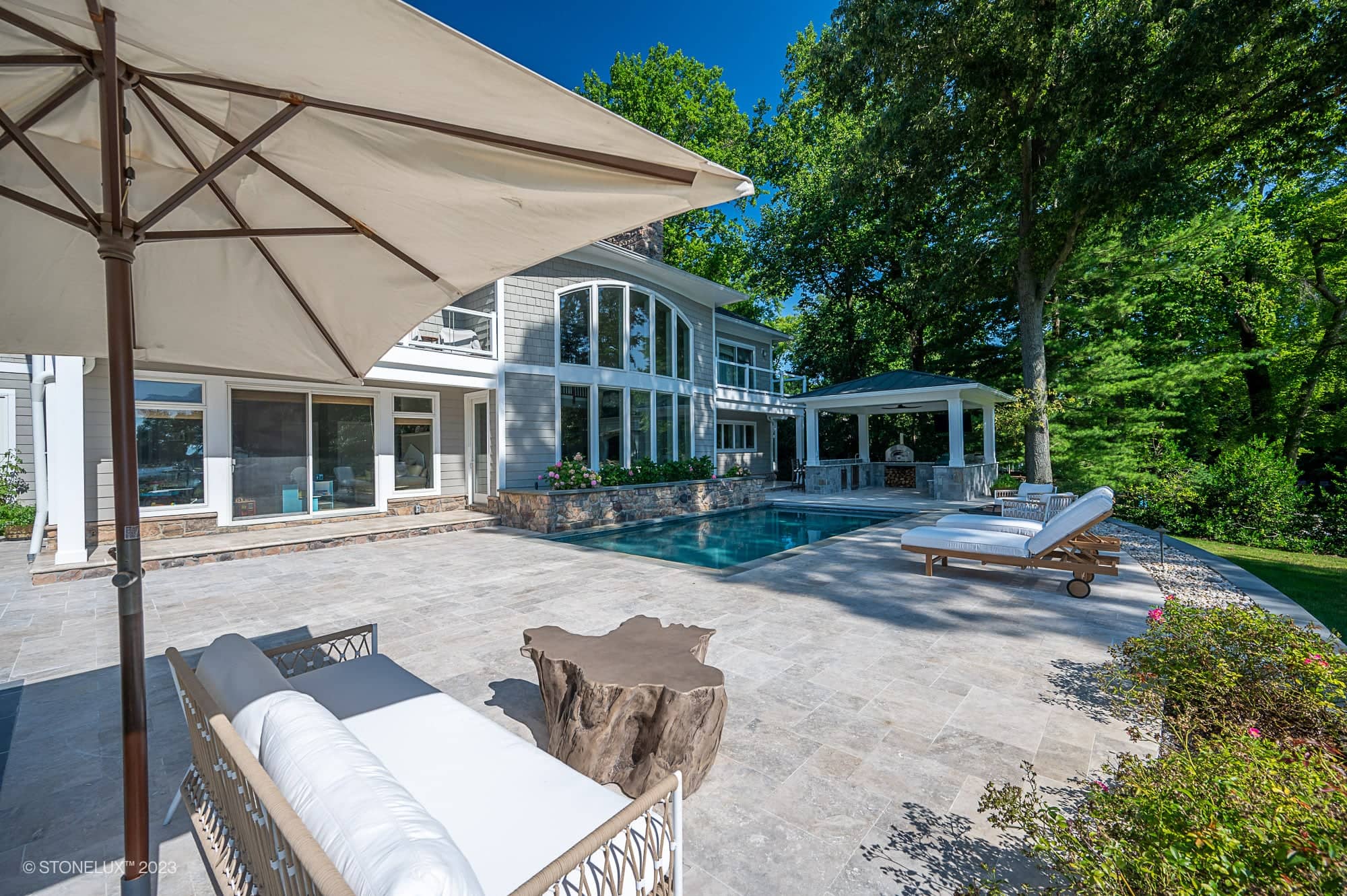 Modern backyard with a swimming pool, sun loungers, and a shaded seating area. The Annapolis Dream – Philly Travertine Pool Project features large windows, surrounded by trees and greenery under a clear blue sky.
