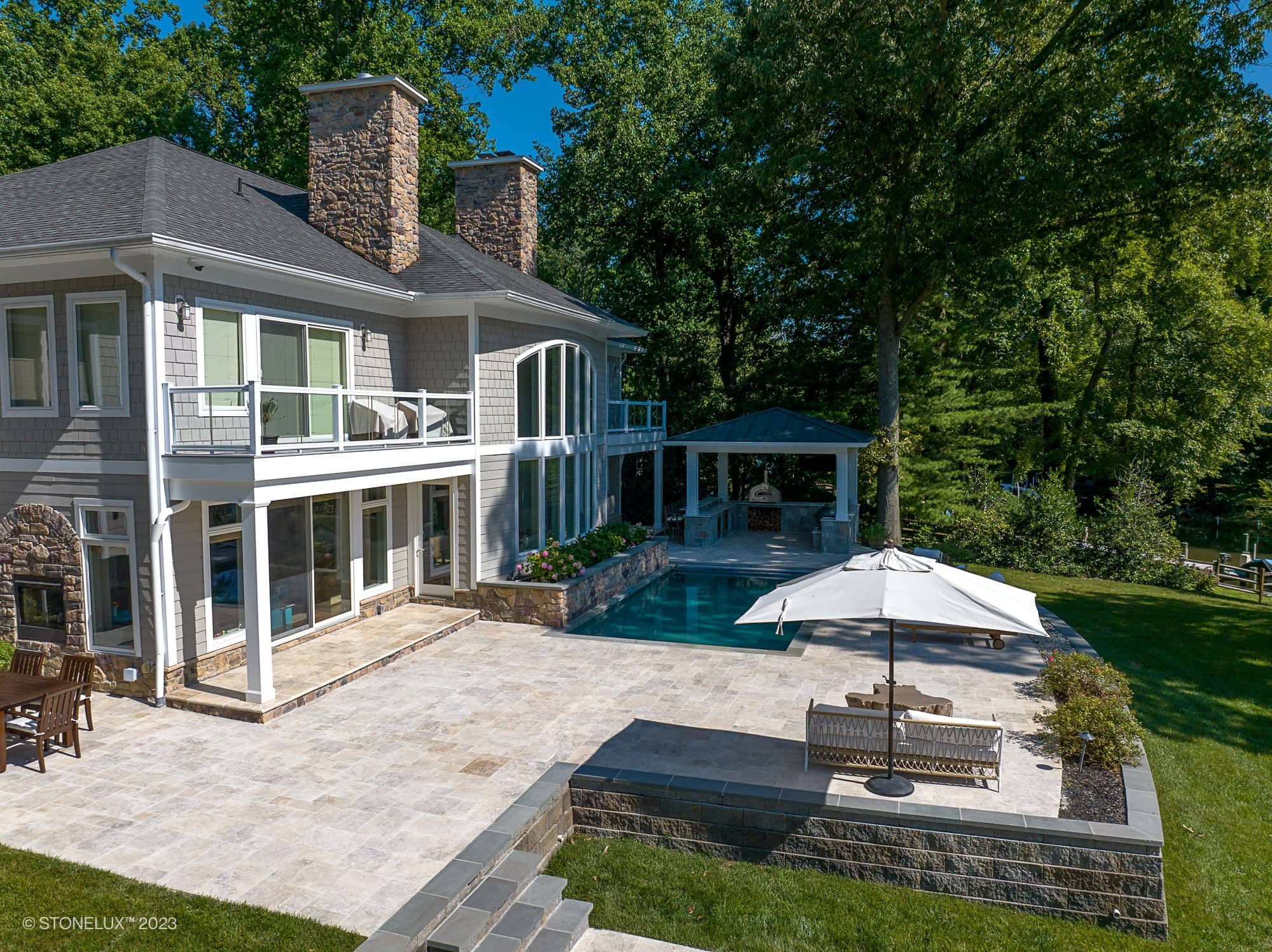 A modern two-story house with large windows, a patio, swimming pool, and pergola features the Annapolis Dream – Philly Travertine Pool Project, with outdoor seating shaded by an umbrella and lush landscaping on a sunny day.