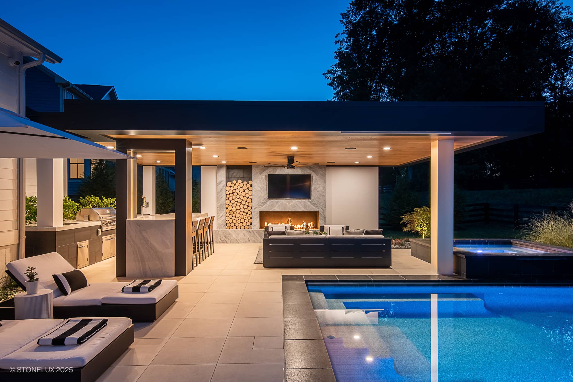 Aldie Modern Escape – Porcelain & Basalt Luxury Pool Pavilion features a modern outdoor patio at dusk with a pool, lounge chairs, open kitchen and bar, TV, stacked firewood, and hot tub. Warm lighting creates a cozy, inviting atmosphere.