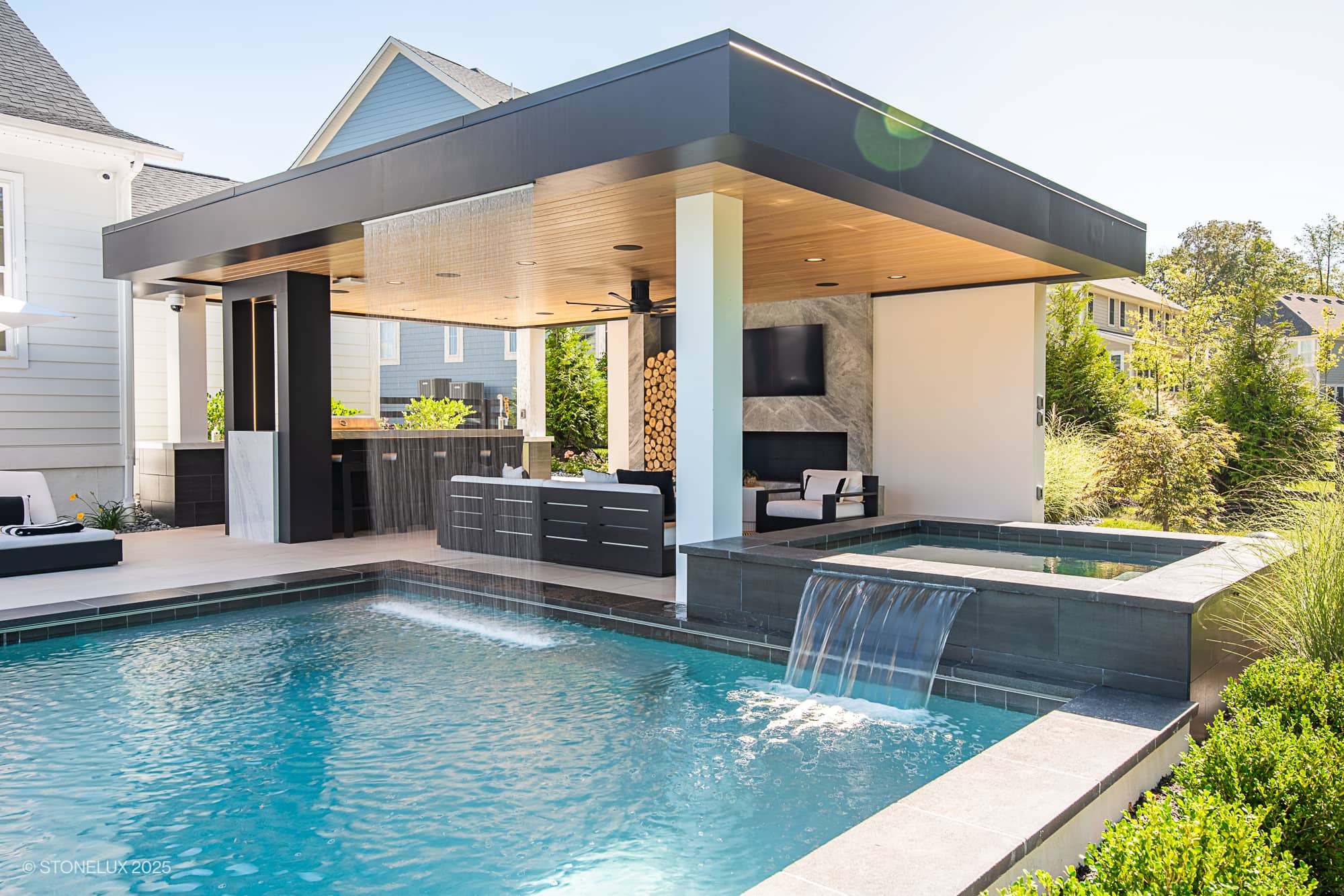 The Aldie Modern Escape – Porcelain & Basalt Luxury Pool Pavilion features a swimming pool with a waterfall, hot tub, and a covered outdoor lounge with TV, kitchen, and bar—all surrounded by greenery and sleek contemporary houses.