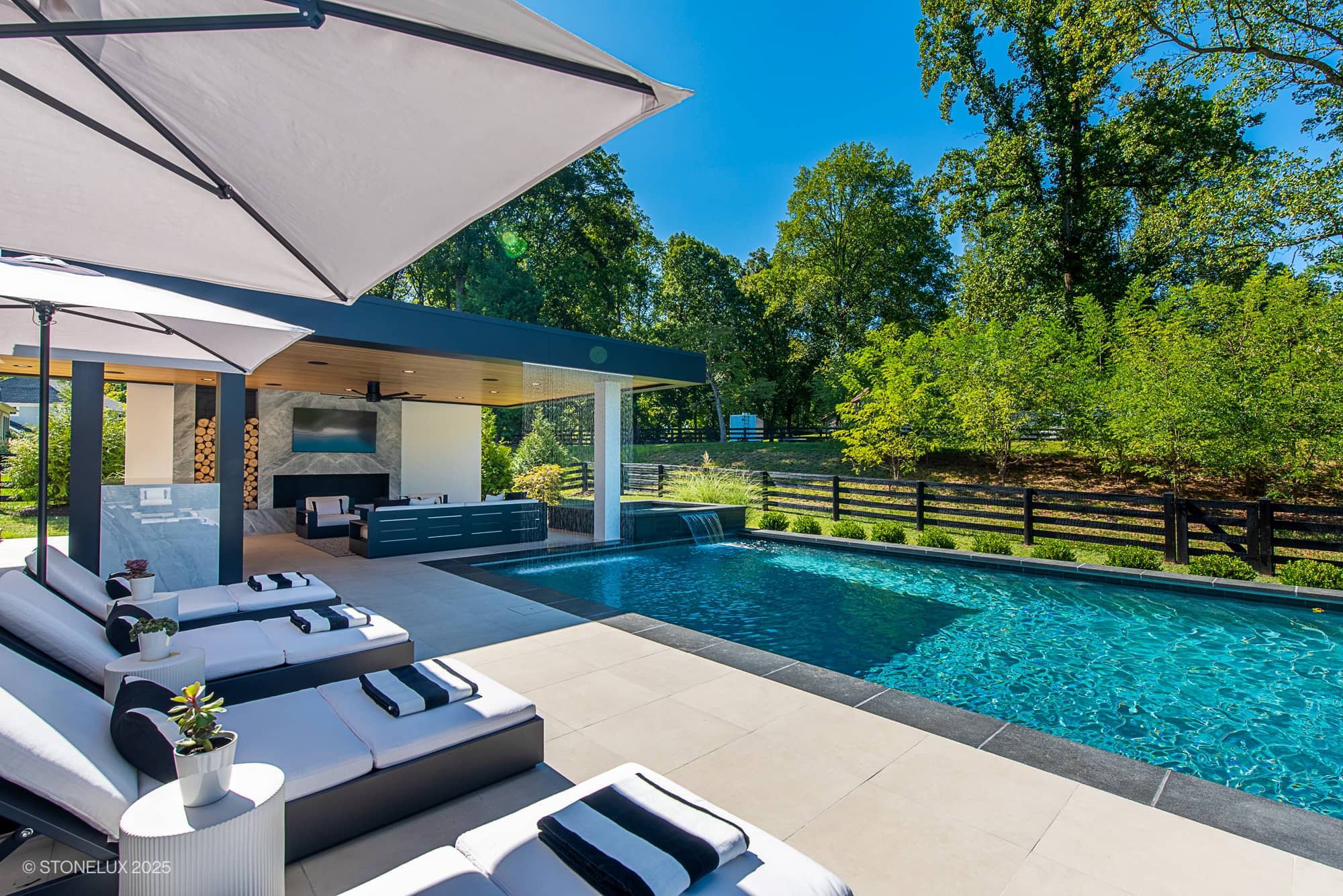 Experience the Aldie Modern Escape – a Porcelain & Basalt Luxury Pool Pavilion featuring lounge chairs, an umbrella, a covered seating space, waterfall feature, and lush green trees framing this modern outdoor oasis on a sunny day.