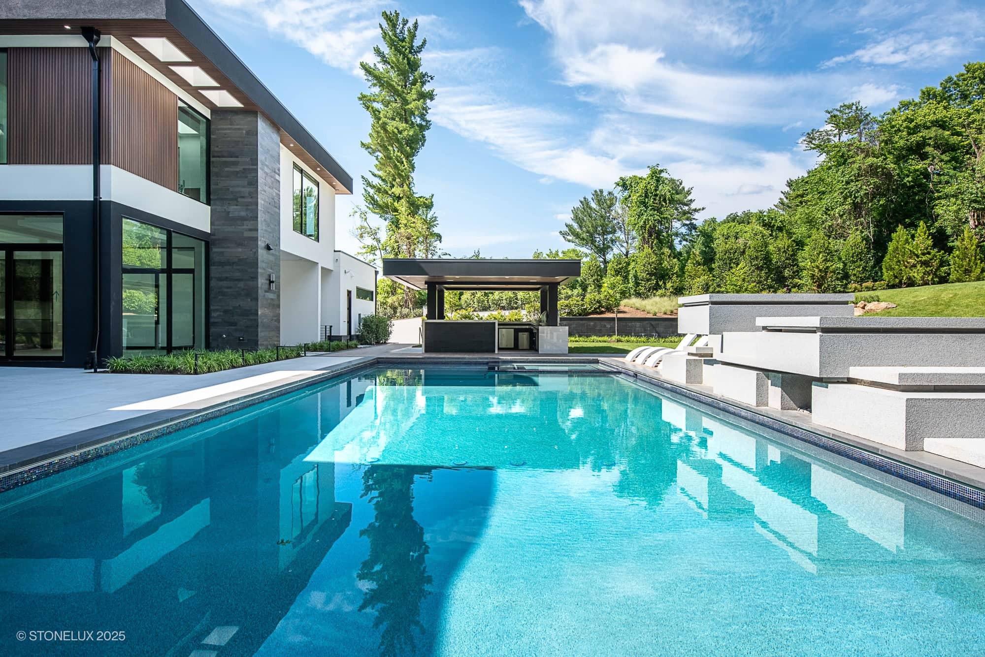 A modern hillside elegance—McLean Retreat—features large windows and wood accents beside a clear blue pool. Enjoy an outdoor kitchen with Overland Pearl Porcelain & Black Basalt, surrounded by trees beneath a bright, partly cloudy sky.