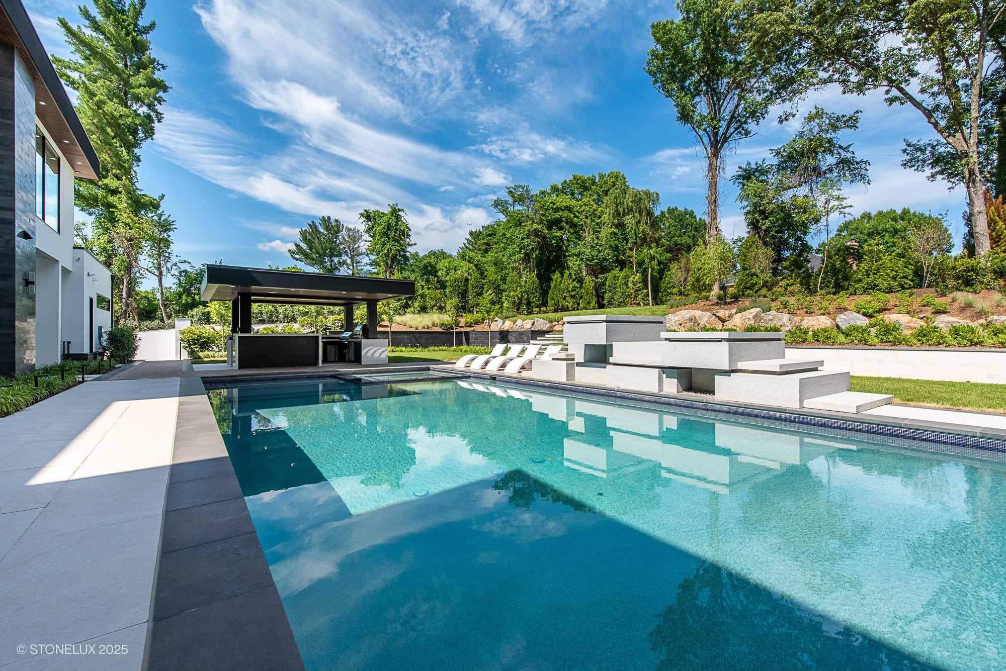 A modern outdoor swimming pool with clear blue water, surrounded by lounge chairs and shady seating areas, showcases Modern Hillside Elegance – McLean Retreat with Overland Pearl Porcelain & Black Basalt accents amid lush green trees.