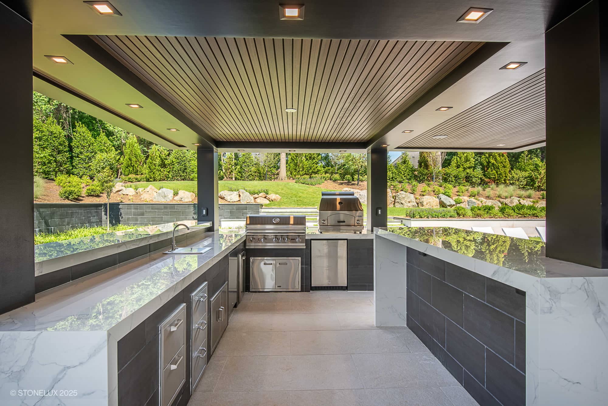 Modern Hillside Elegance defines this outdoor kitchen, featuring Overland Pearl Porcelain, black basalt details, marble countertops, and stainless steel appliances. Surrounded by greenery, the covered patio offers built-in grills.