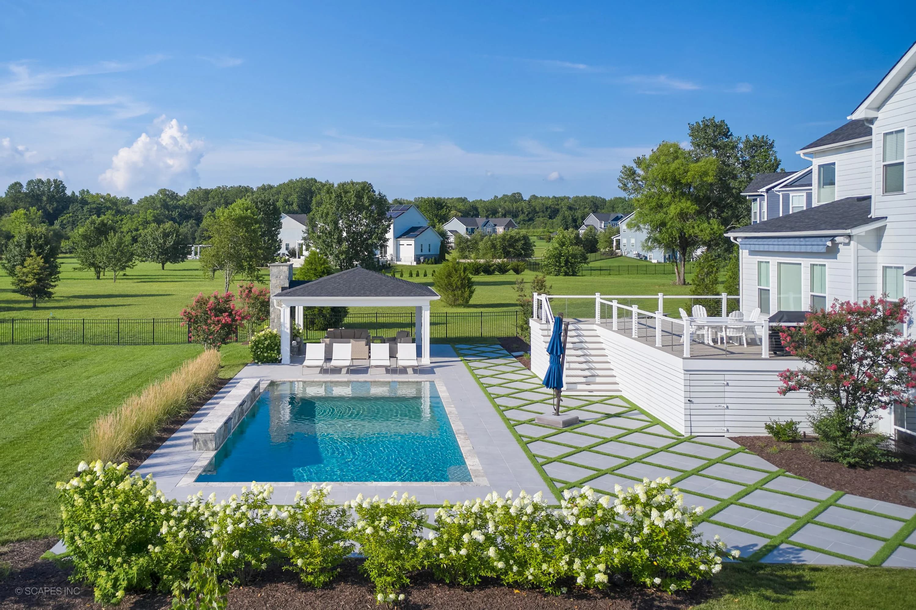 Modern backyard in Annapolis, MD featuring Golf Course Geometry design with Porcelain Pavers and Silver Travertine from STONELUX. Sleek pool with clear blue water, surrounded by lush greenery, white deck chairs, and a stylish pergola.