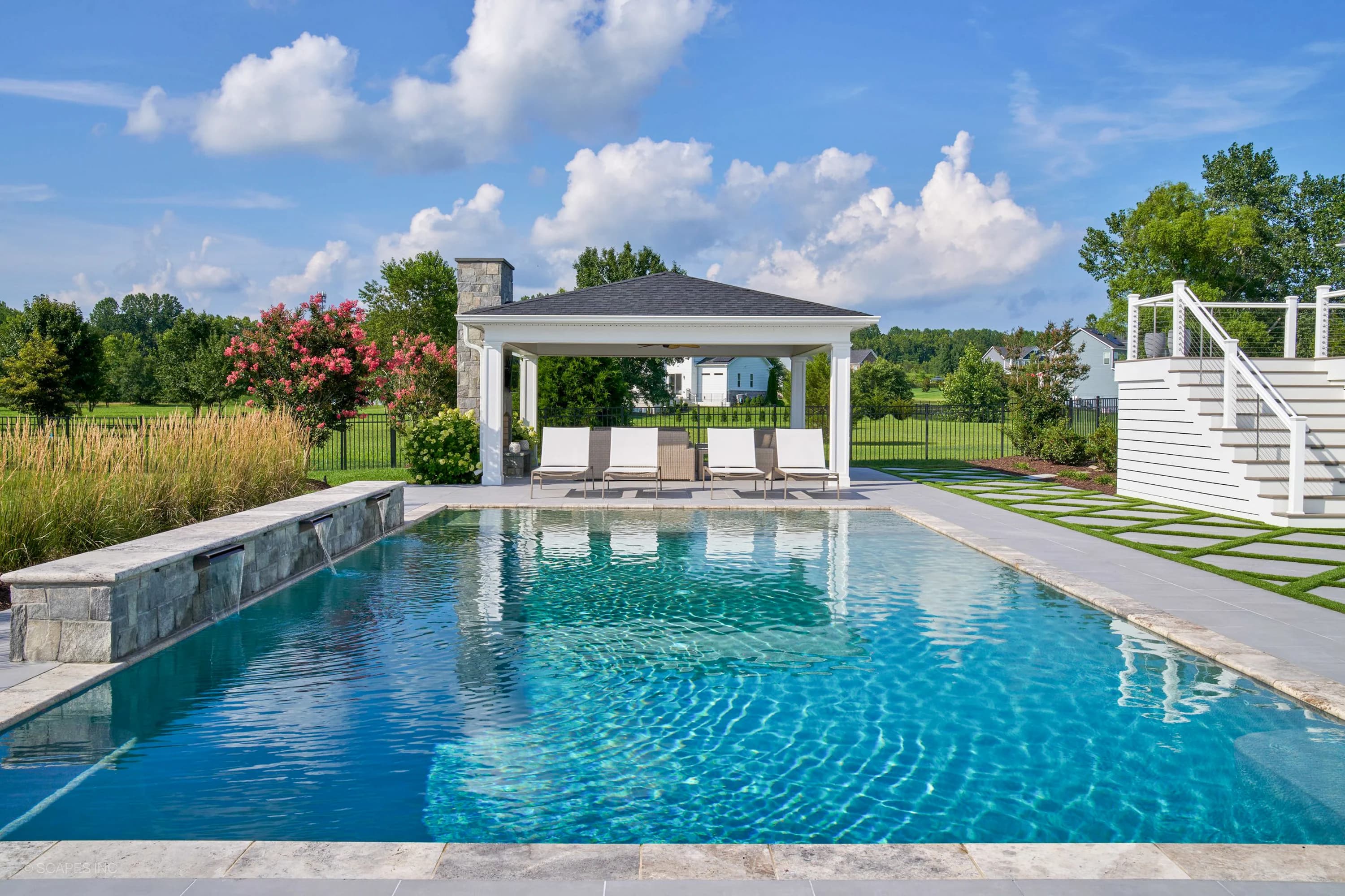 Modern pool area in Annapolis, MD, featuring Golf Course Geometry: Porcelain Pavers with Turf from the Silver Travertine collection. Blue pool water is surrounded by grey pavers, a pavilion, white lounge chairs, and lush landscaping, creating a serene ...