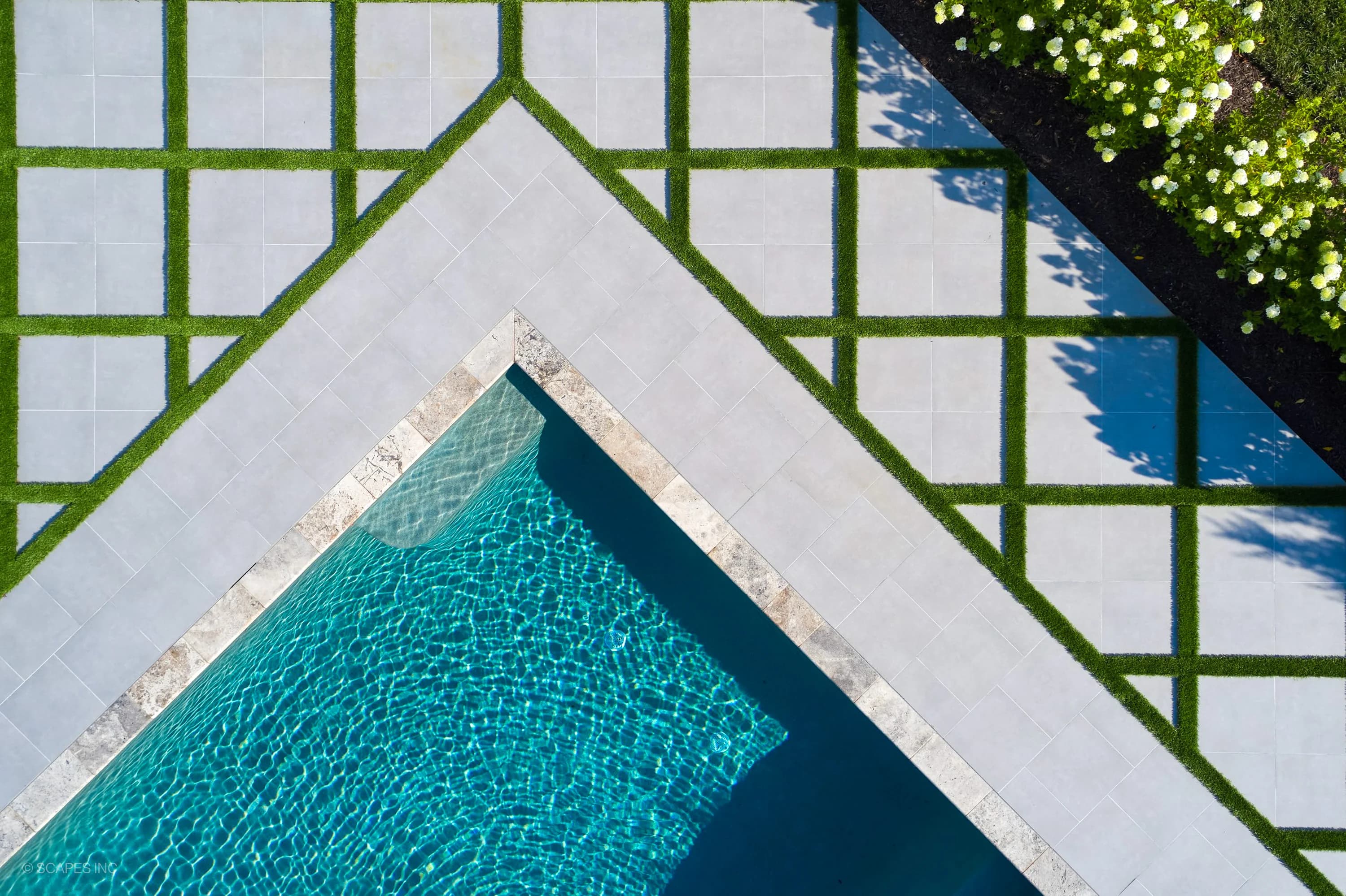 Aerial view of a modern hardscape patio with STONELUX Porcelain Pavers from the Silver Travertine collection, featuring geometric turf patterns. The sparkling blue pool complements the sleek pavers, surrounded by lush green landscaping in Annapolis, MD.