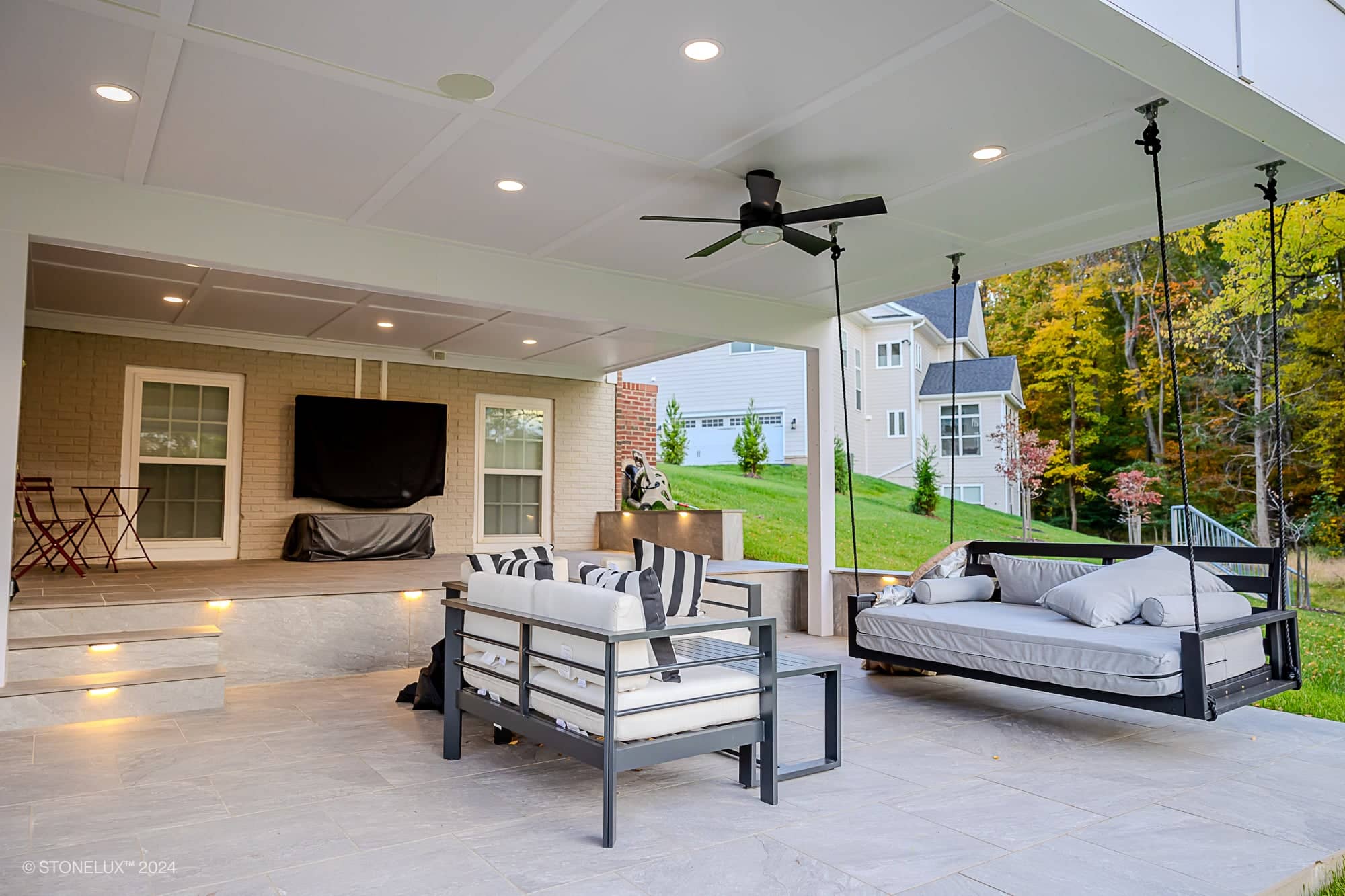 A modern covered patio with a ceiling fan, cushioned seating, swing bed, and outdoor TV features a Rasa Grey: Porcelain Paver Patio in Fairfax. Steps lead to a grassy yard, with house exteriors and fall foliage in the background.