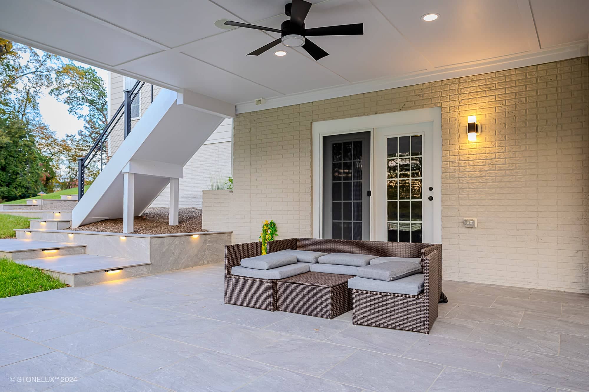 A modern Rasa Grey: Porcelain Paver Patio in Fairfax features a covered patio with a wicker sectional sofa and gray cushions, ceiling fan, wall light, stairs leading up, French doors to the house, and greenery and lawn in the background.