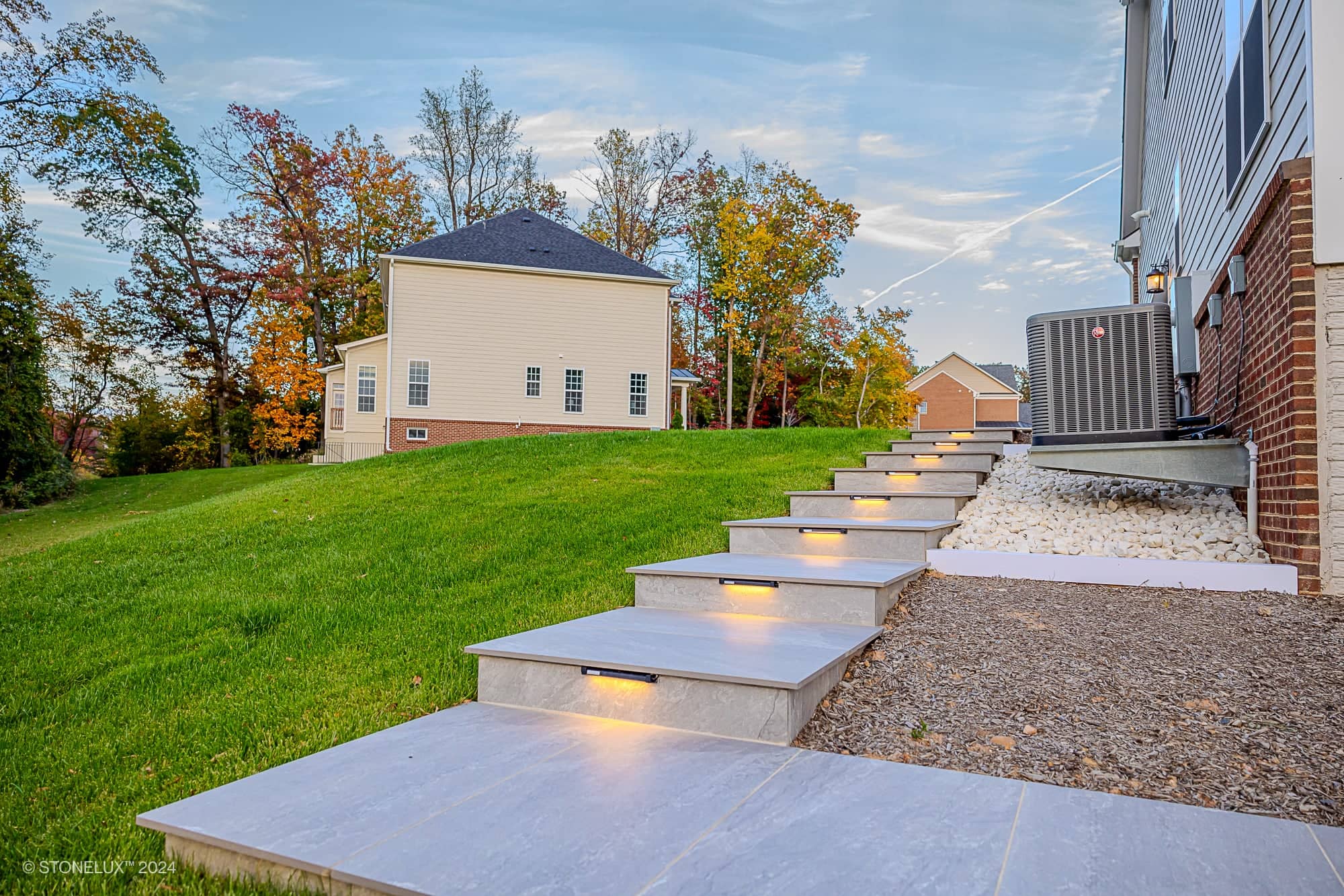 Modern outdoor stone steps with built-in lights lead up a grassy hillside between two houses, complemented by a Rasa Grey: Porcelain Paver Patio in Fairfax, with trees in the background showing early autumn colors.