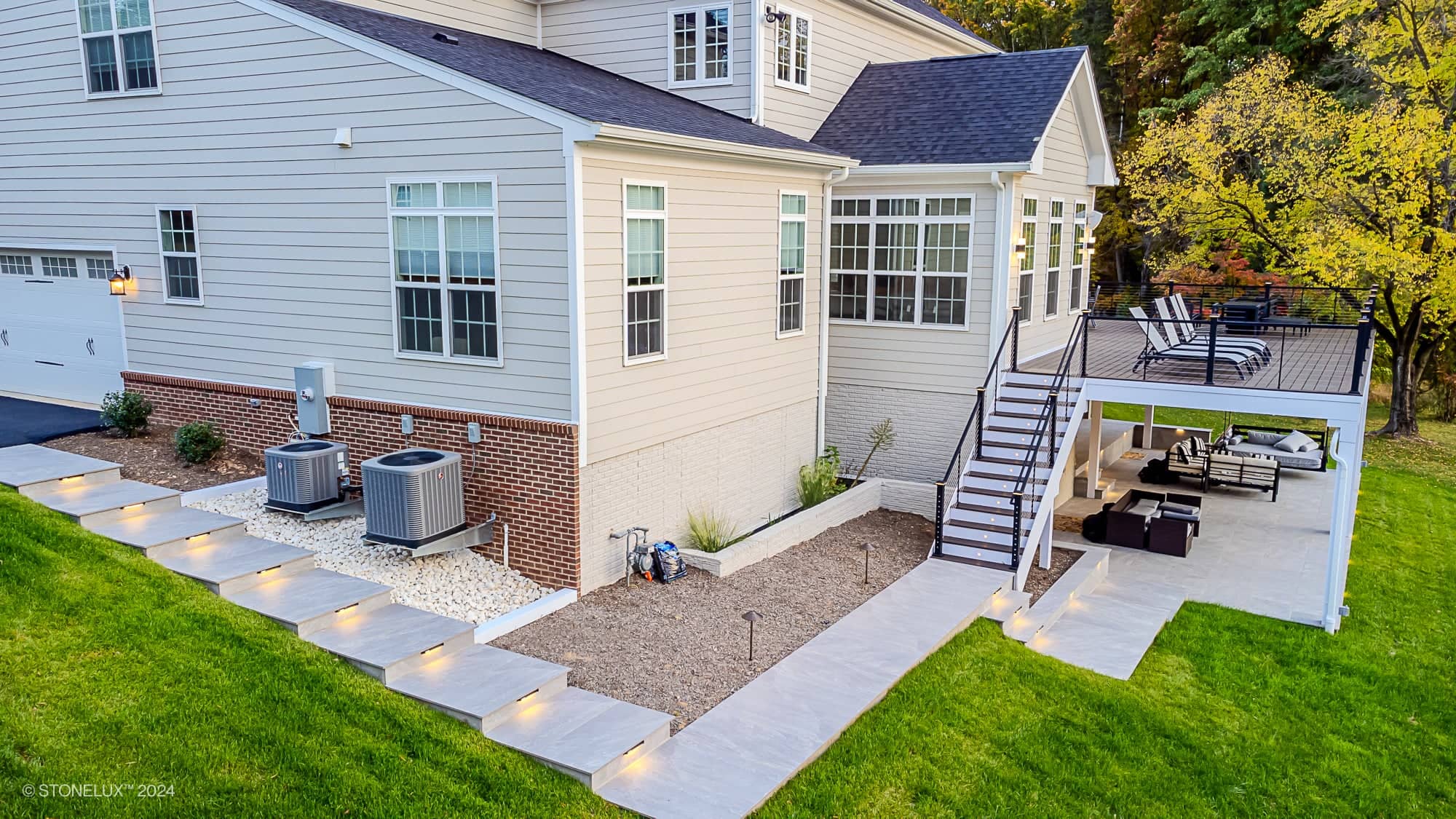 A modern house with a spacious Rasa Grey: Porcelain Paver Patio in Fairfax, upper deck with lounge chairs, and lower seating area. Concrete steps with lights lead to the landscaped yard featuring stones and two HVAC units.