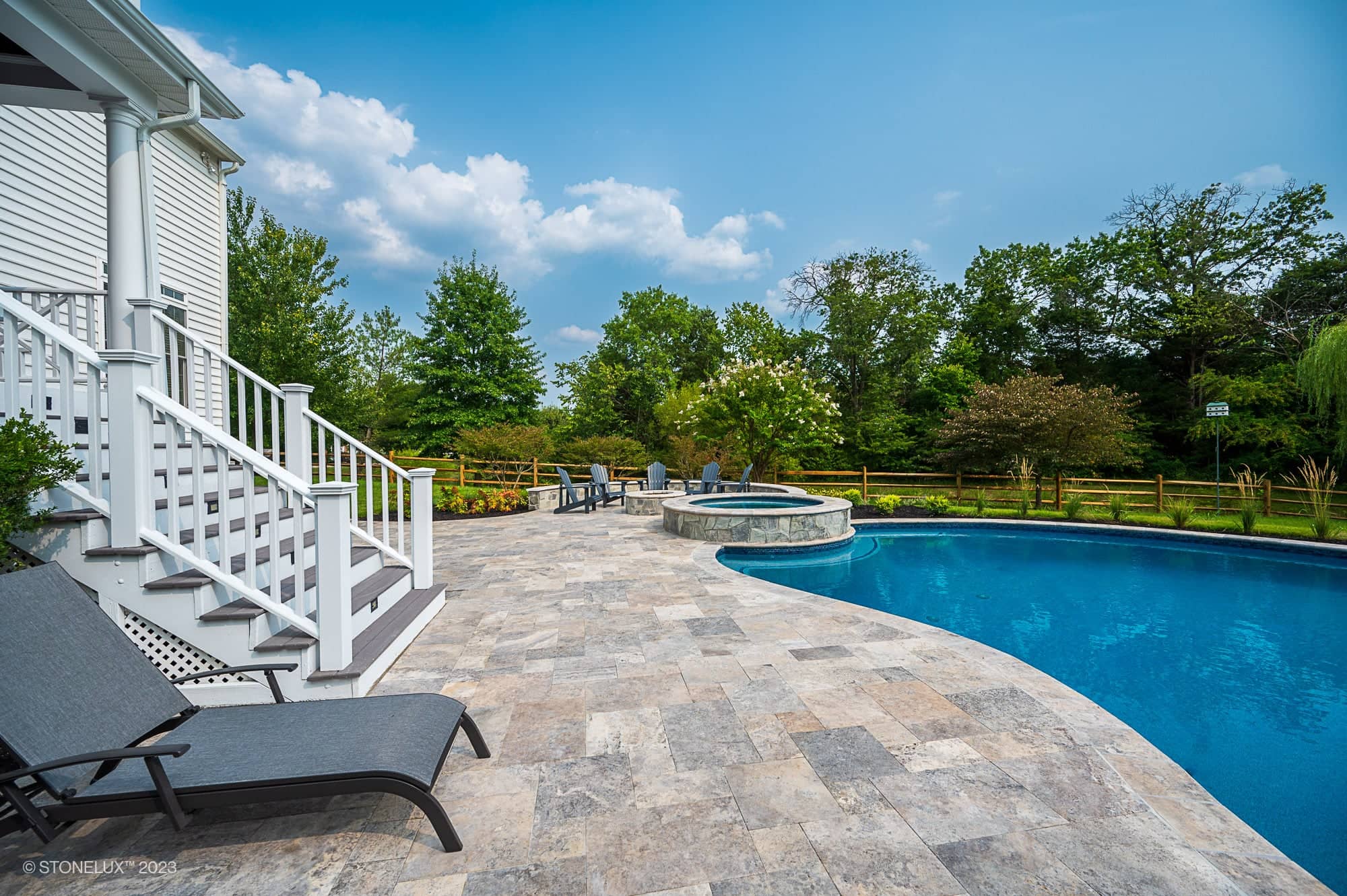 A backyard oasis featuring Silver Splash: The Silver Travertine Pool Haven with a curved swimming pool, hot tub, and lounge chairs. White house stairs descend to the stone-tiled deck, framed by green trees and a wooden fence under a partly cloudy sky.