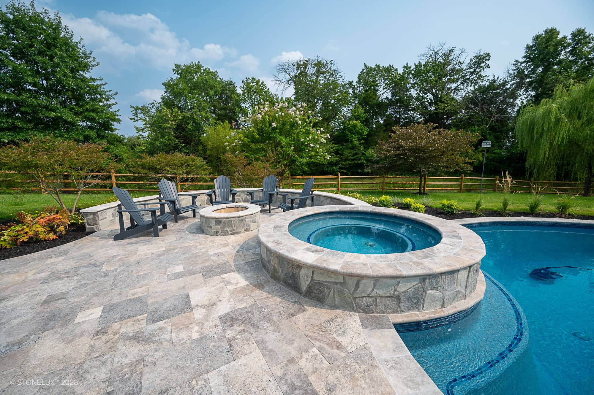 A backyard pool, Silver Splash: The Silver Travertine Pool Haven, features a stone hot tub and a paved patio. Adirondack chairs circle a stone fire pit, all framed by green trees and a wooden fence under a blue sky.