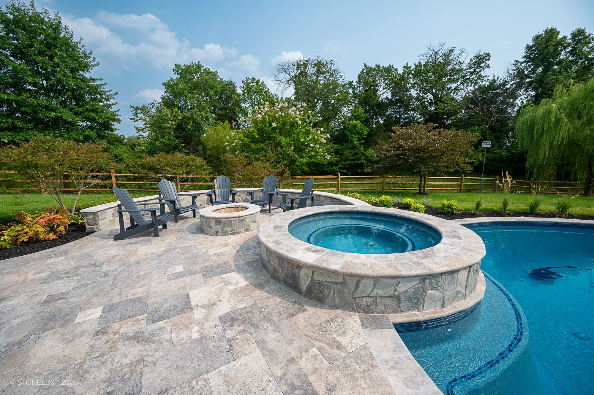 A backyard pool, Silver Splash: The Silver Travertine Pool Haven, features a stone hot tub and a paved patio. Adirondack chairs circle a stone fire pit, all framed by green trees and a wooden fence under a blue sky.