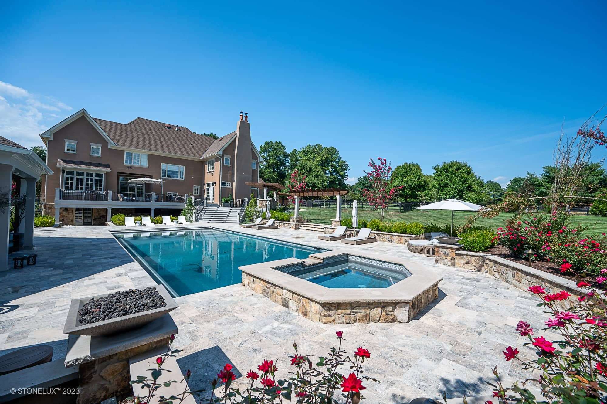 Luxurious backyard featuring the Silver Expanse: The Silver Travertine Pool Landscape, with a pool, hot tub, fire pit, lounge chairs, manicured landscaping, and a large house in the background under a clear blue sky.