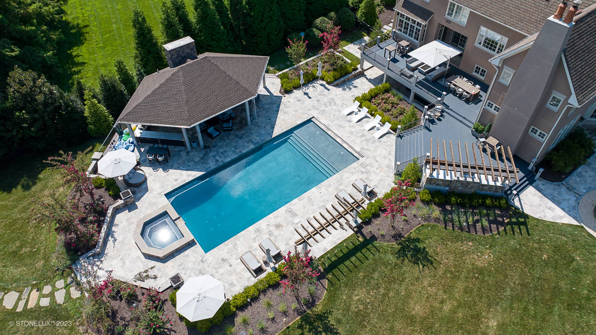 Aerial view of a large backyard featuring the Silver Expanse: The Silver Travertine Pool Landscape, with a rectangular swimming pool, attached hot tub, poolside lounge chairs, shaded seating areas, and landscaped gardens beside a spacious house.