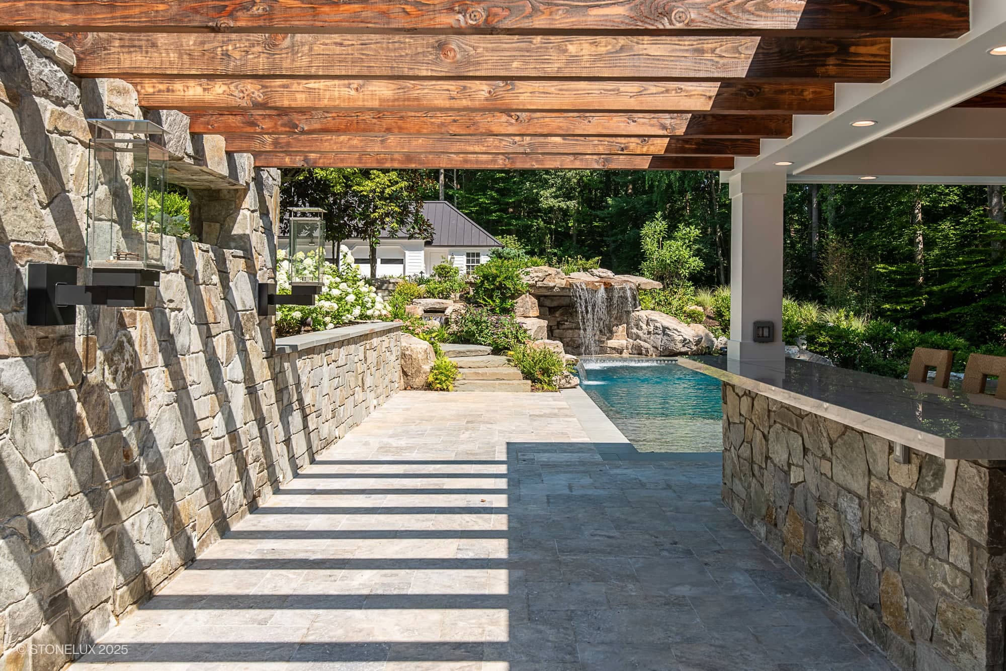 The Clifton Cascade Retreat – Silver Travertine Oasis features a stone patio with a wooden pergola, stone bar, and pool. Sunlight casts shadows on the tiled floor while lush greenery and a small waterfall create a serene backdrop.