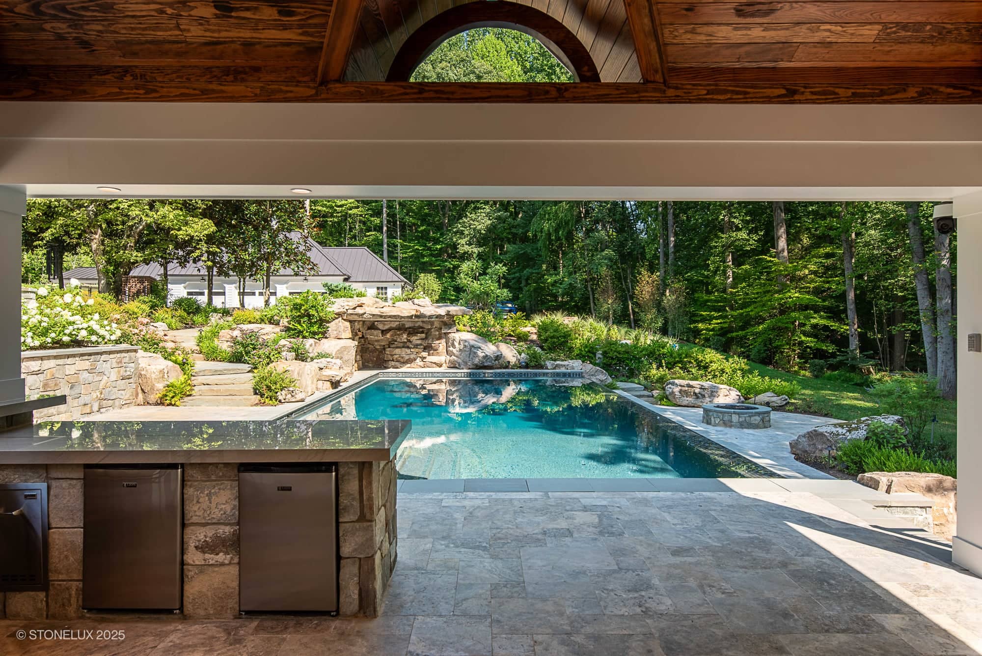 A view from a covered patio with a stone counter and built-in appliances, overlooking the Clifton Cascade Retreat – Silver Travertine Oasis swimming pool, surrounded by lush greenery, flower beds, trees, and a charming house.