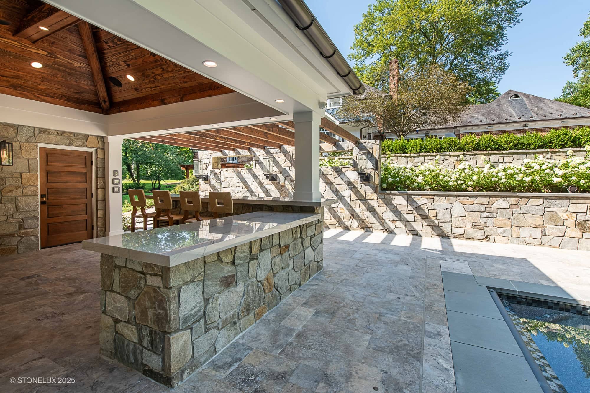 Covered outdoor kitchen and bar area with stone counters, modern stools, and wooden ceiling beside a landscaped patio at Clifton Cascade Retreat – Silver Travertine Oasis, featuring stone walls, white blooms, and a view of the grassy yard and trees.