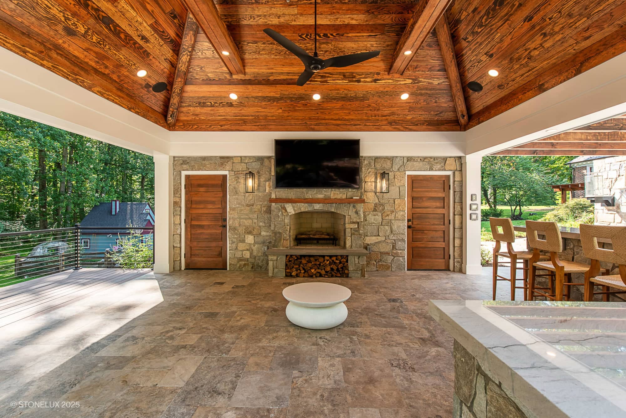 Spacious covered patio at Clifton Cascade Retreat – Silver Travertine Oasis features a wooden vaulted ceiling, stone fireplace, wall-mounted TV, bar seating, and a tiled floor—all overlooking a lush green backyard.