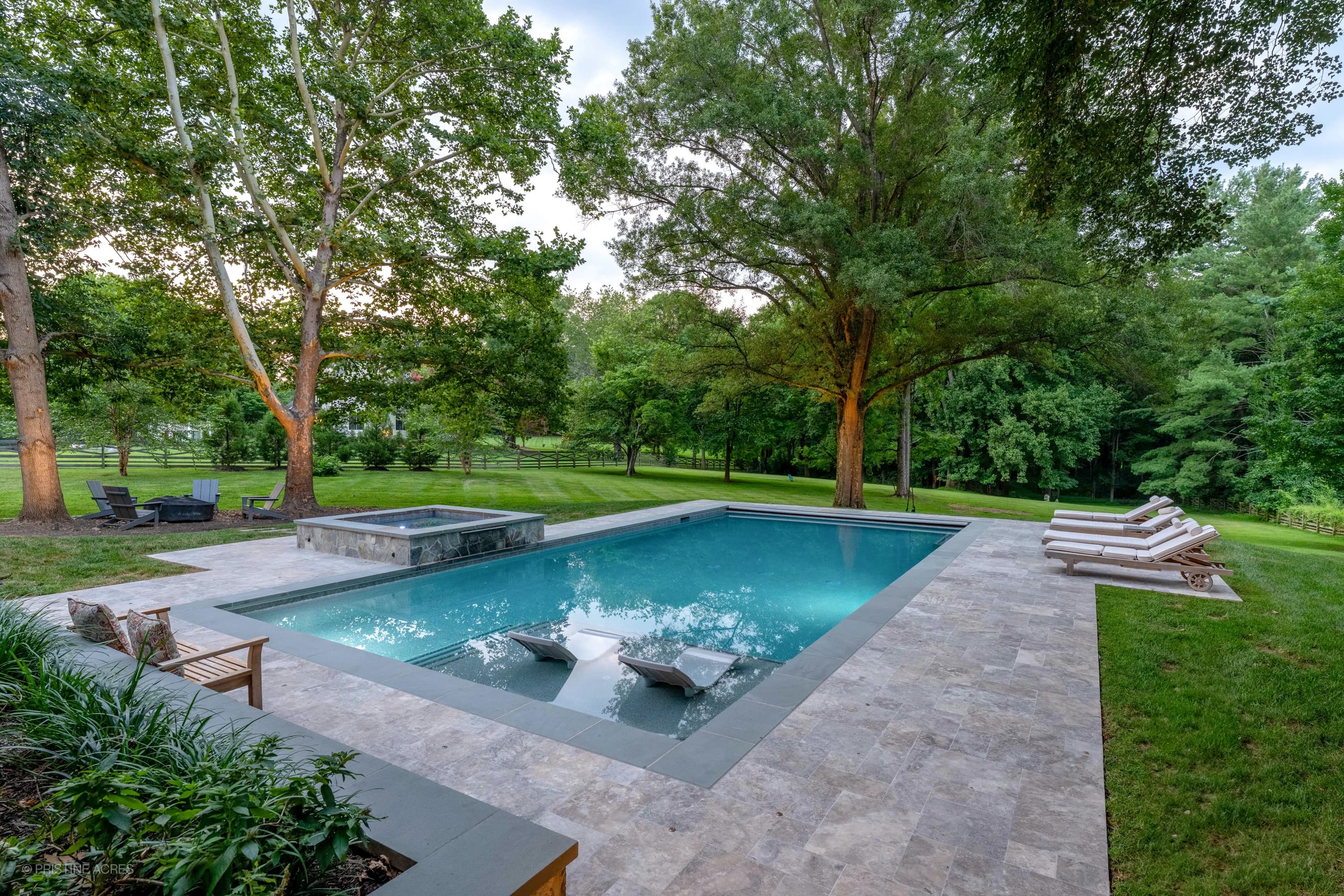 Vienna Pool Retreat in Vienna, VA showcases Silver Travertine pavers with a contemporary design. The elegant pool features grey bluestone coping, complementing the clear blue water. Surrounded by lush greenery, white lounge chairs, and a serene backyar...