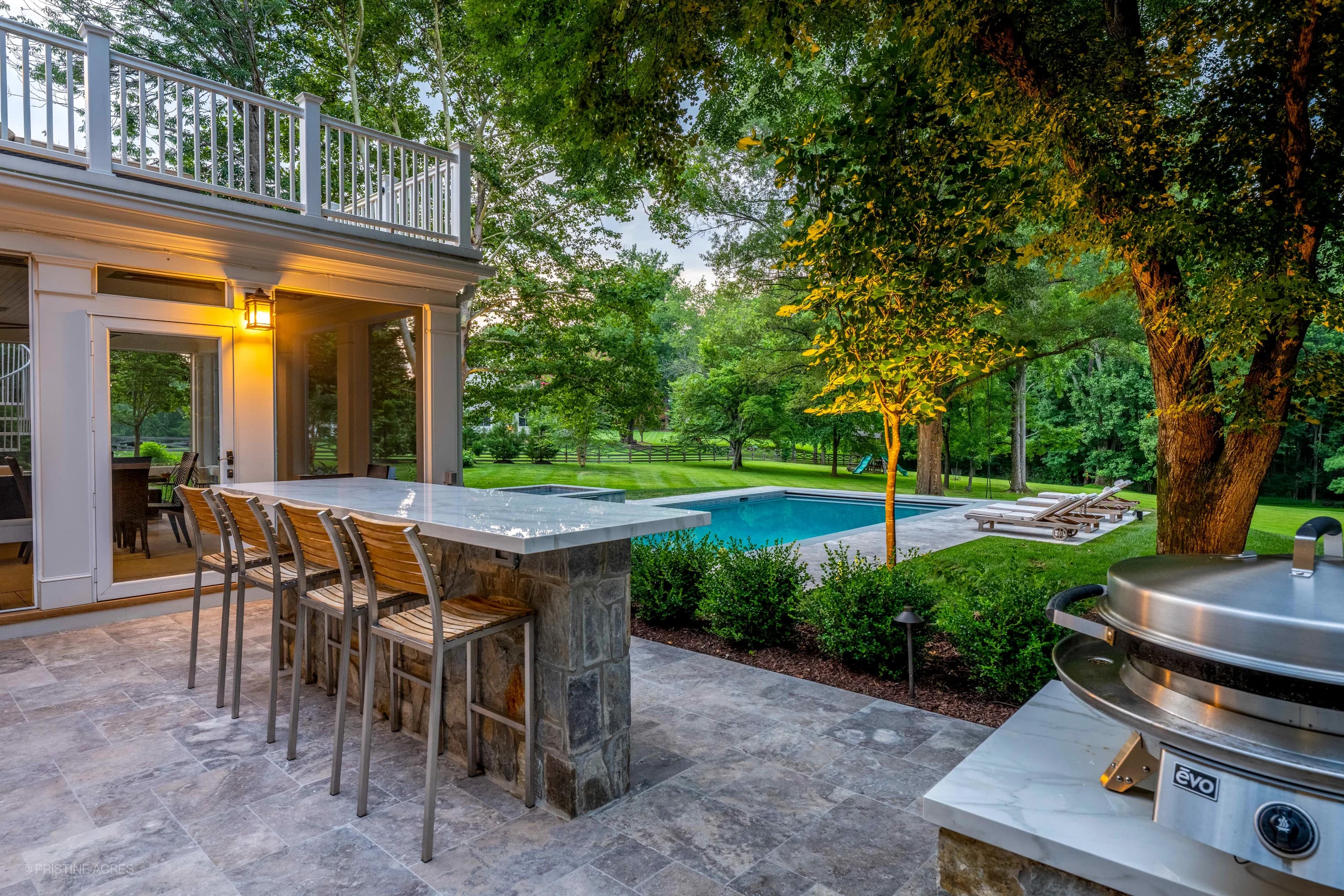 Vienna Pool Retreat in Vienna, VA, features Silver Travertine pavers and Bluestone accents. The modern design showcases a serene blue pool, surrounded by lush greenery and white lounge chairs. A stone bar with seating overlooks the elegant patio.