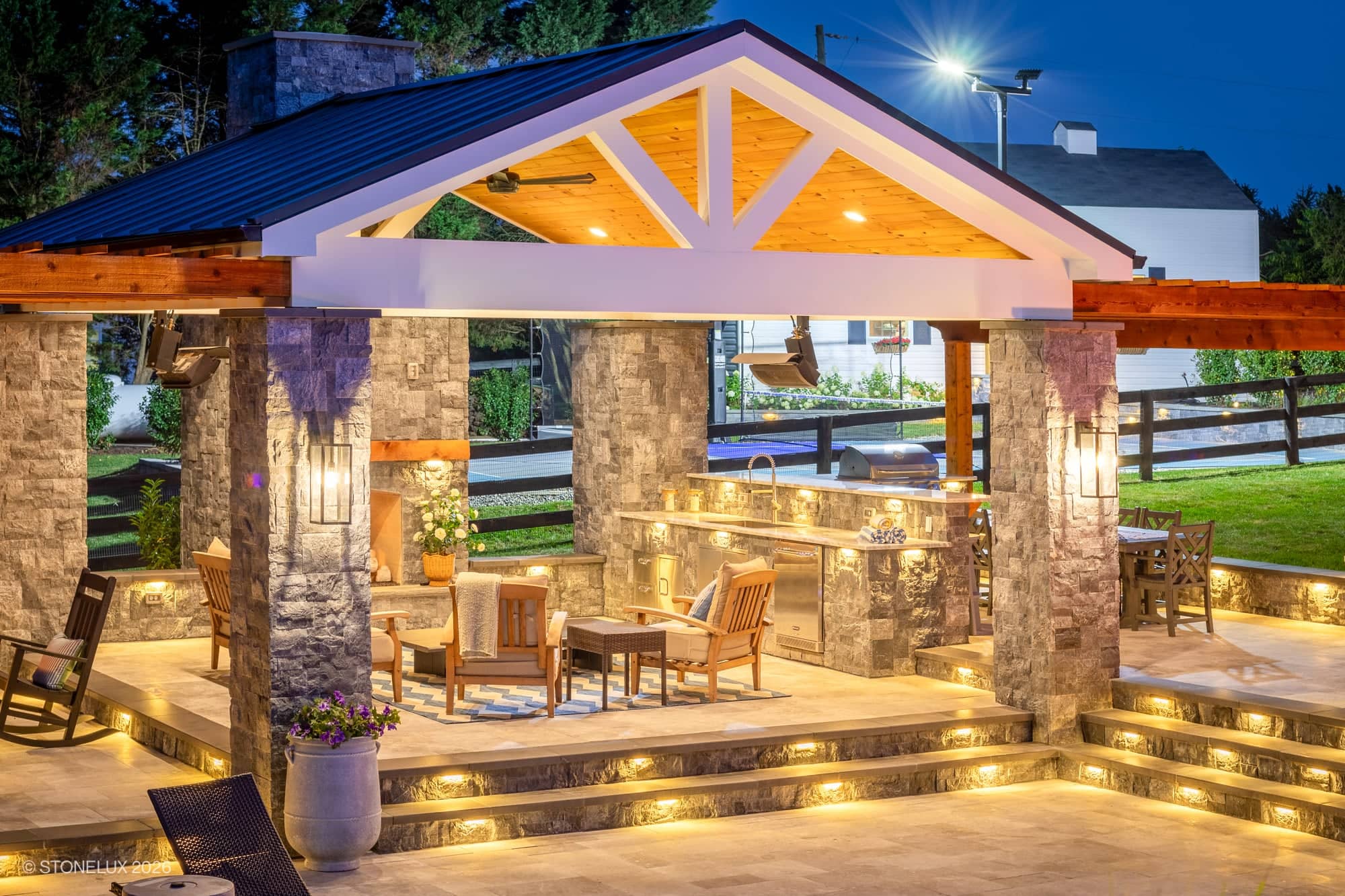 Backyard oasis in McLean, VA featuring Tundra Grey Marble and Black Basalt from Stonelux. Modern outdoor kitchen with Afyon White Marble accents, illuminated steps, wooden pergola, cozy seating, lush landscaping, and ambient evening lighting.