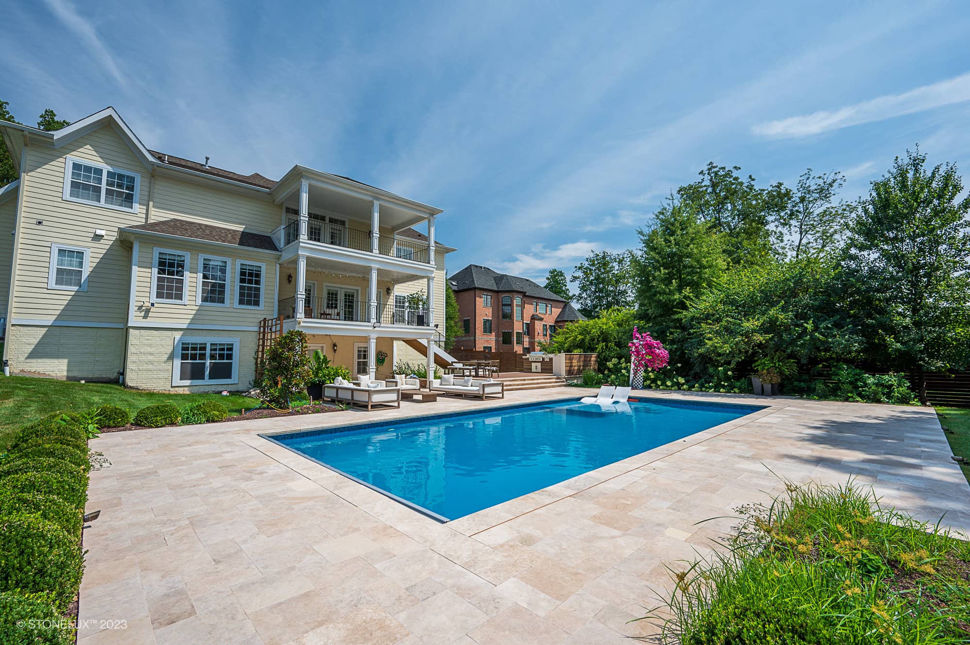 A spacious backyard features a rectangular swimming pool surrounded by Brown Serenity: The Walnut Travertine Oasis stone tiles, lounge chairs, and neatly trimmed shrubs. A large, light-colored house with multiple balconies overlooks the pool area.