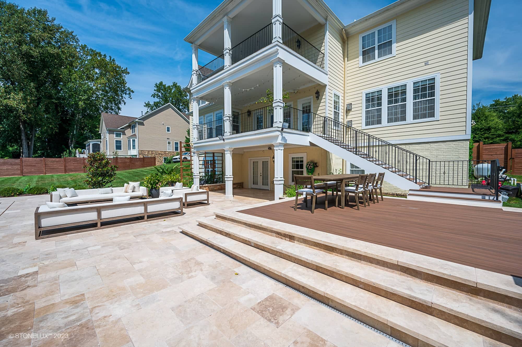 A spacious backyard patio with Brown Serenity: The Walnut Travertine Oasis beige tiles features an outdoor lounge area with white cushioned sofas, a dining table on a wooden deck, and a large, multi-story yellow house in the background.