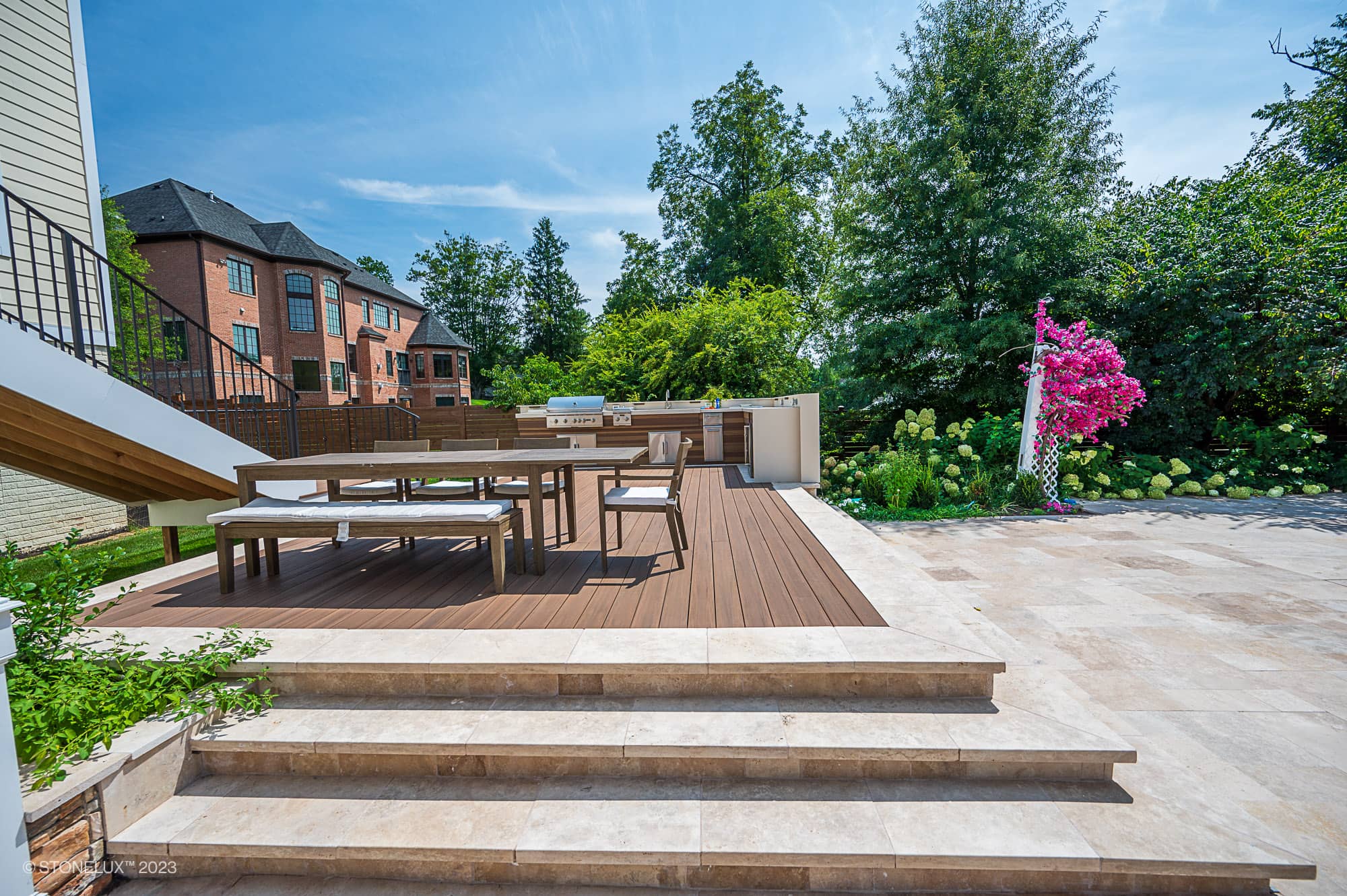 A modern outdoor patio with wooden decking features picnic-style tables and benches, a built-in grill, steps leading down to a tiled area in Brown Serenity: The Walnut Travertine Oasis, and lush greenery with flowering plants surrounding the space.