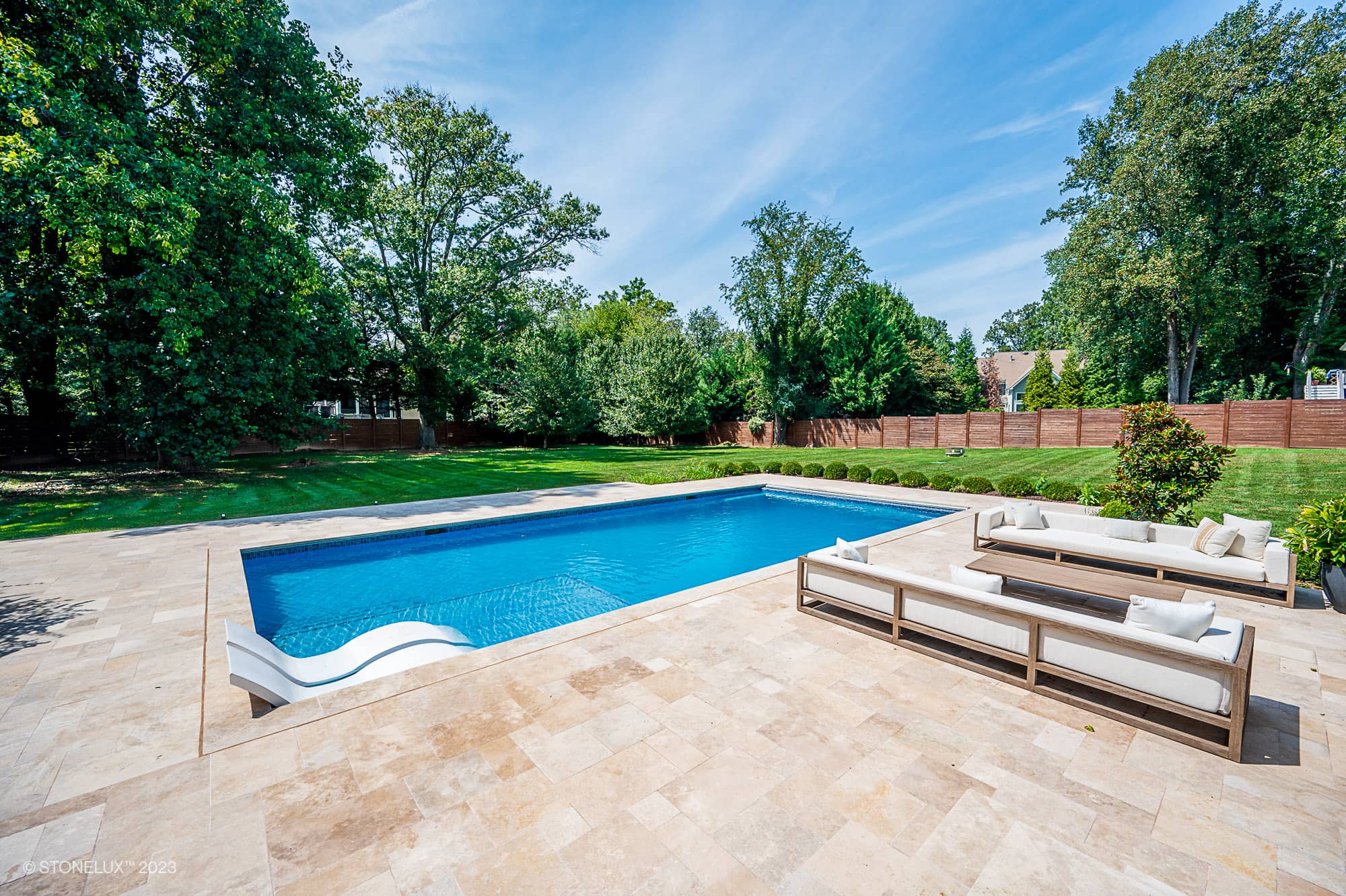 A modern backyard with a rectangular swimming pool, a white pool slide, and beige outdoor sofas on a stone patio. Experience Brown Serenity: The Walnut Travertine Oasis, surrounded by green grass, trees, and a wooden privacy fence under a blue sky.