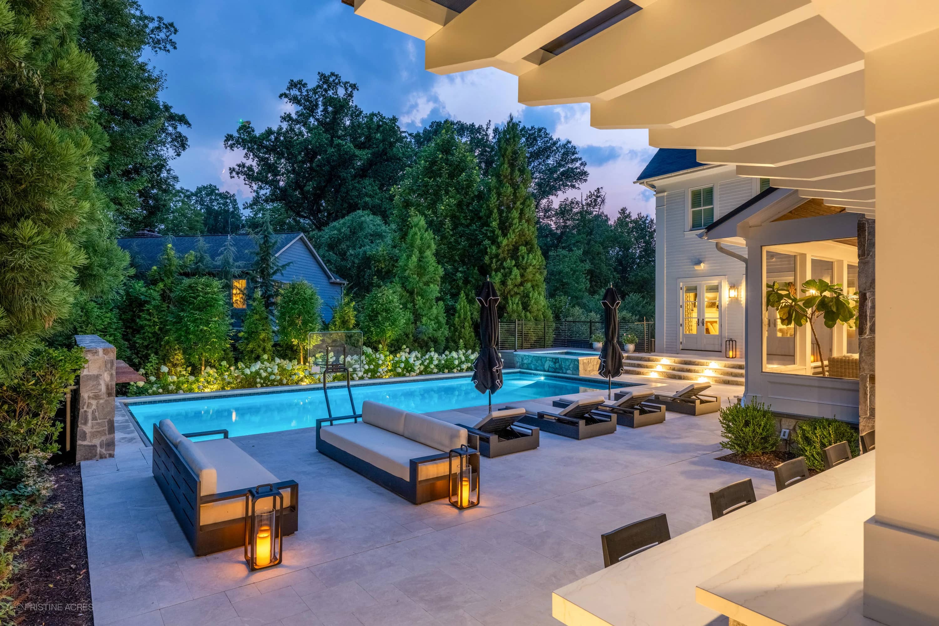 Luxurious outdoor living space in McLean, VA featuring Pera Cream Marble. Modern design with sleek pool, illuminated blue water, elegant lounge chairs, and lush landscaping. Marble pavers harmonize with ambient lighting and contemporary architecture.
