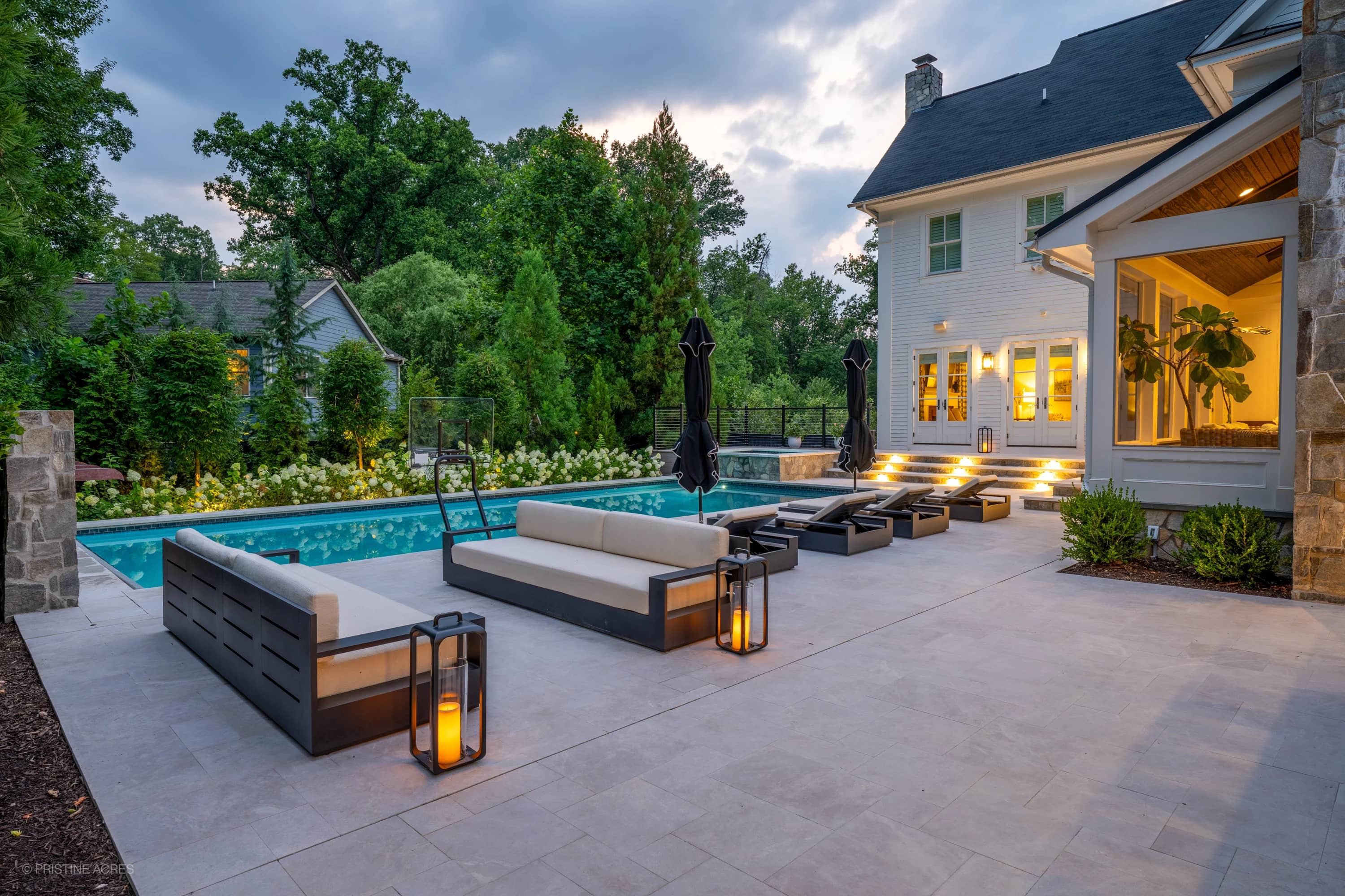 Luxurious outdoor living space in McLean, VA featuring Pera Cream Marble pavers surrounding a serene blue pool. Modern design with sleek black loungers, soft lighting, and lush greenery. Ideal for hardscape and pool renovation projects.