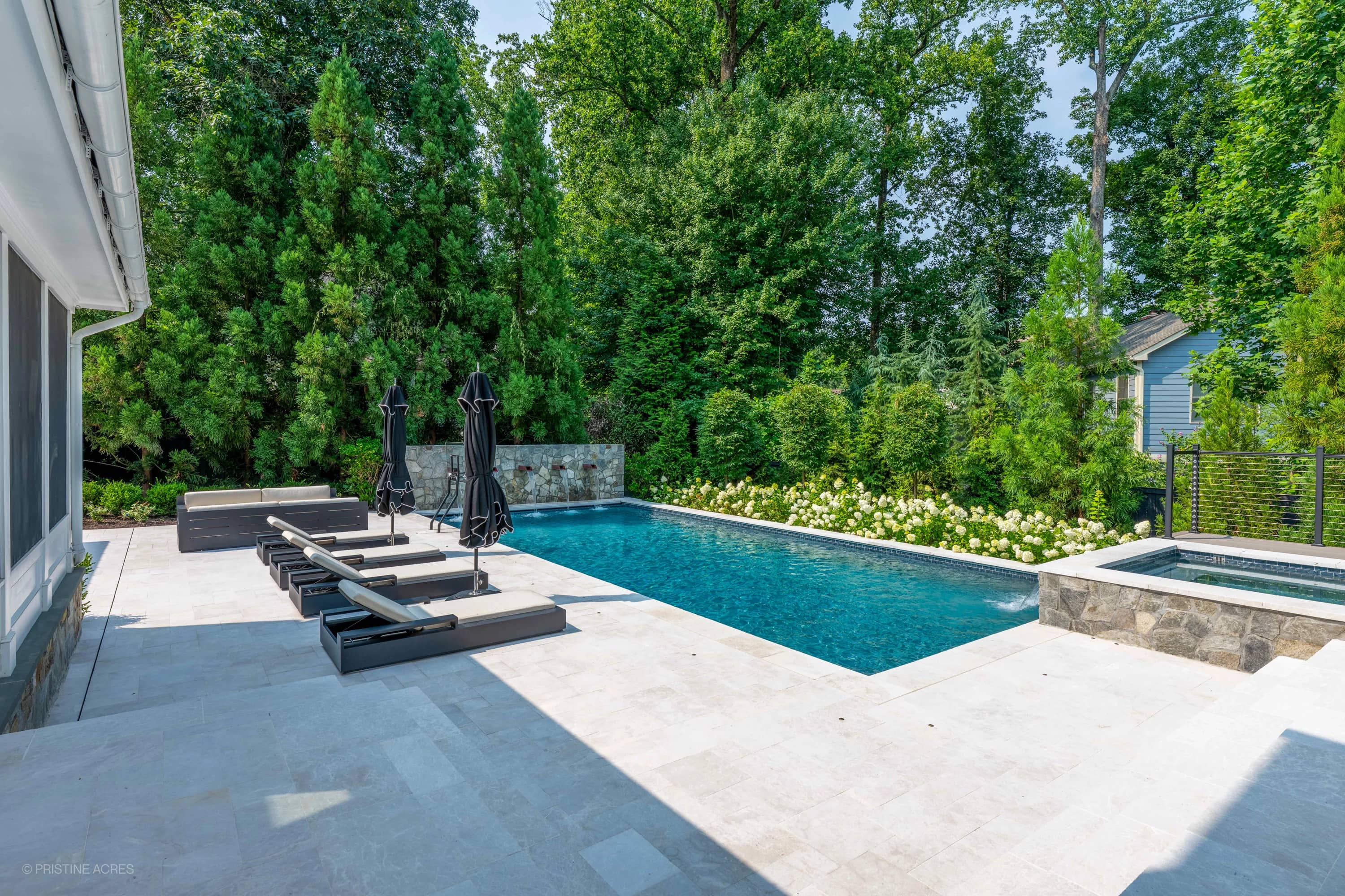 Modern outdoor living space featuring Pera Cream Marble pavers surrounding a deep blue pool in McLean, VA. The contemporary design includes sleek grey lounge chairs, black umbrellas, lush greenery, and elegant stonework, creating a serene backyard oasis.
