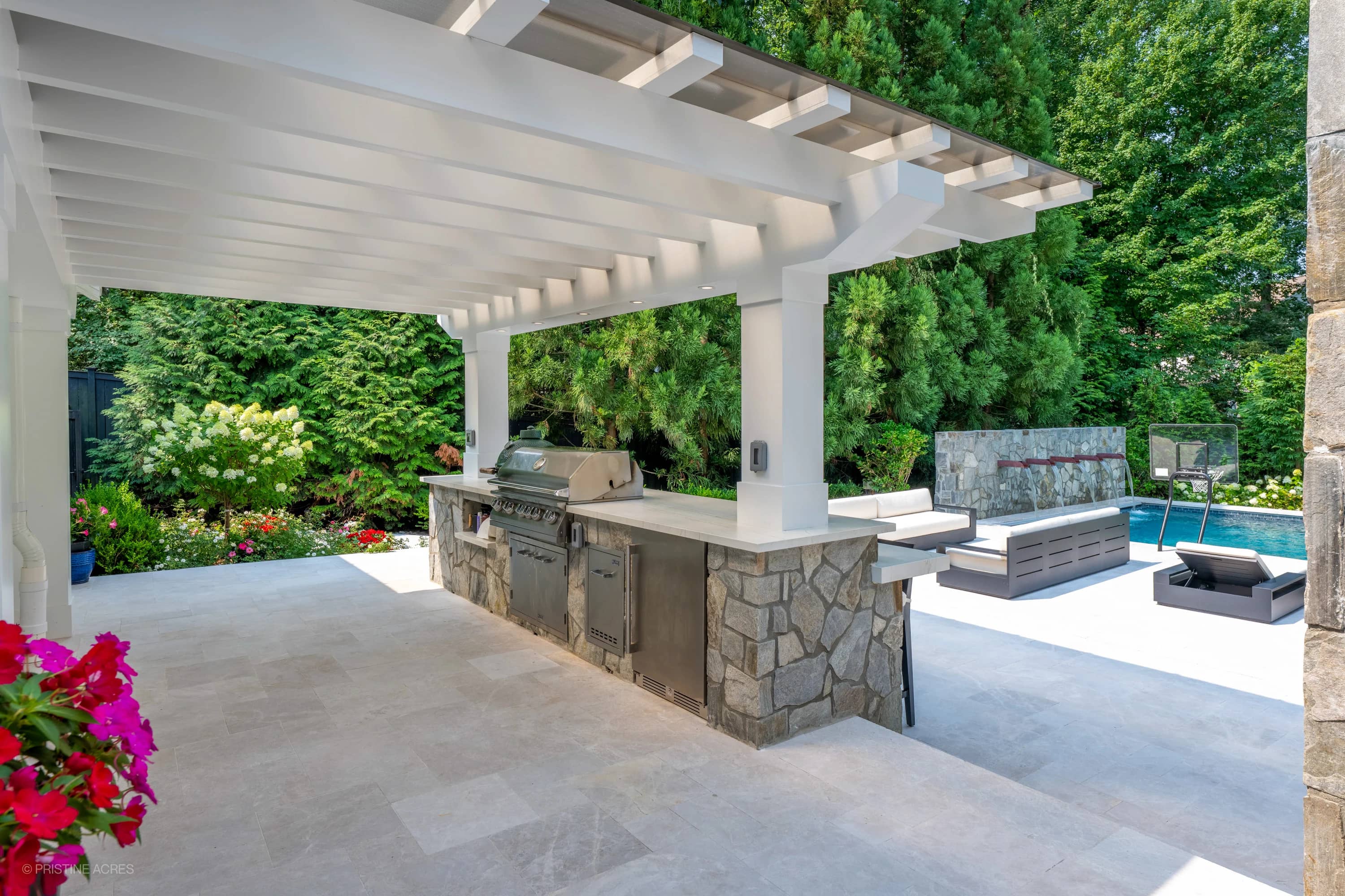 Outdoor living space in McLean, VA, featuring Pera Cream Marble flooring. The modern design includes a stainless steel grill under a pergola, surrounded by lush greenery. Adjacent is a pool with blue water, stone accents, and contemporary seating, crea...
