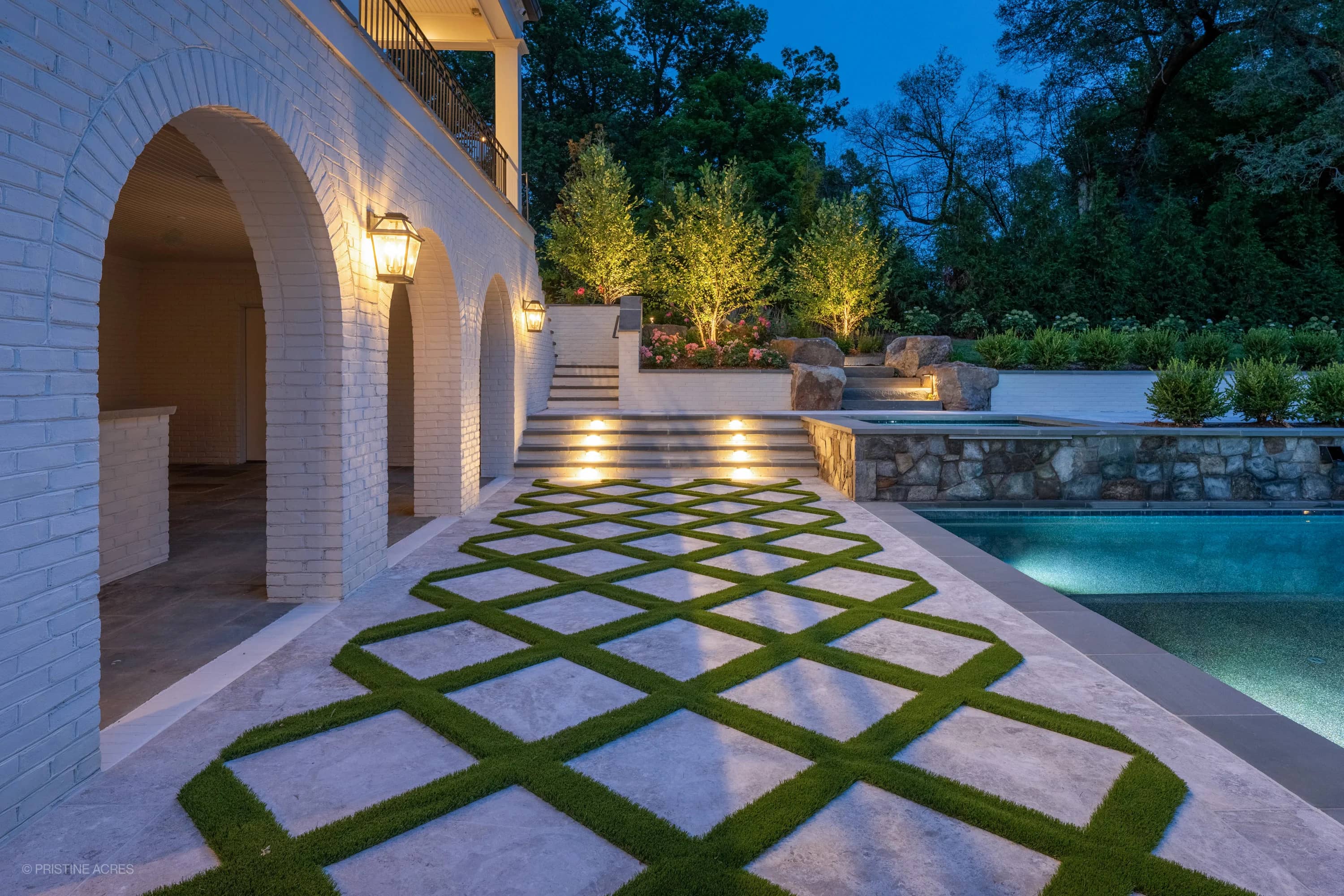 Tundra Grey Marble pool deck with Bluestone coping in McLean, VA. Features elegant stone pavers with lush greenery accents, a shimmering blue pool, and warm outdoor lighting. Modern architectural style with landscaped surroundings enhances the luxuriou...