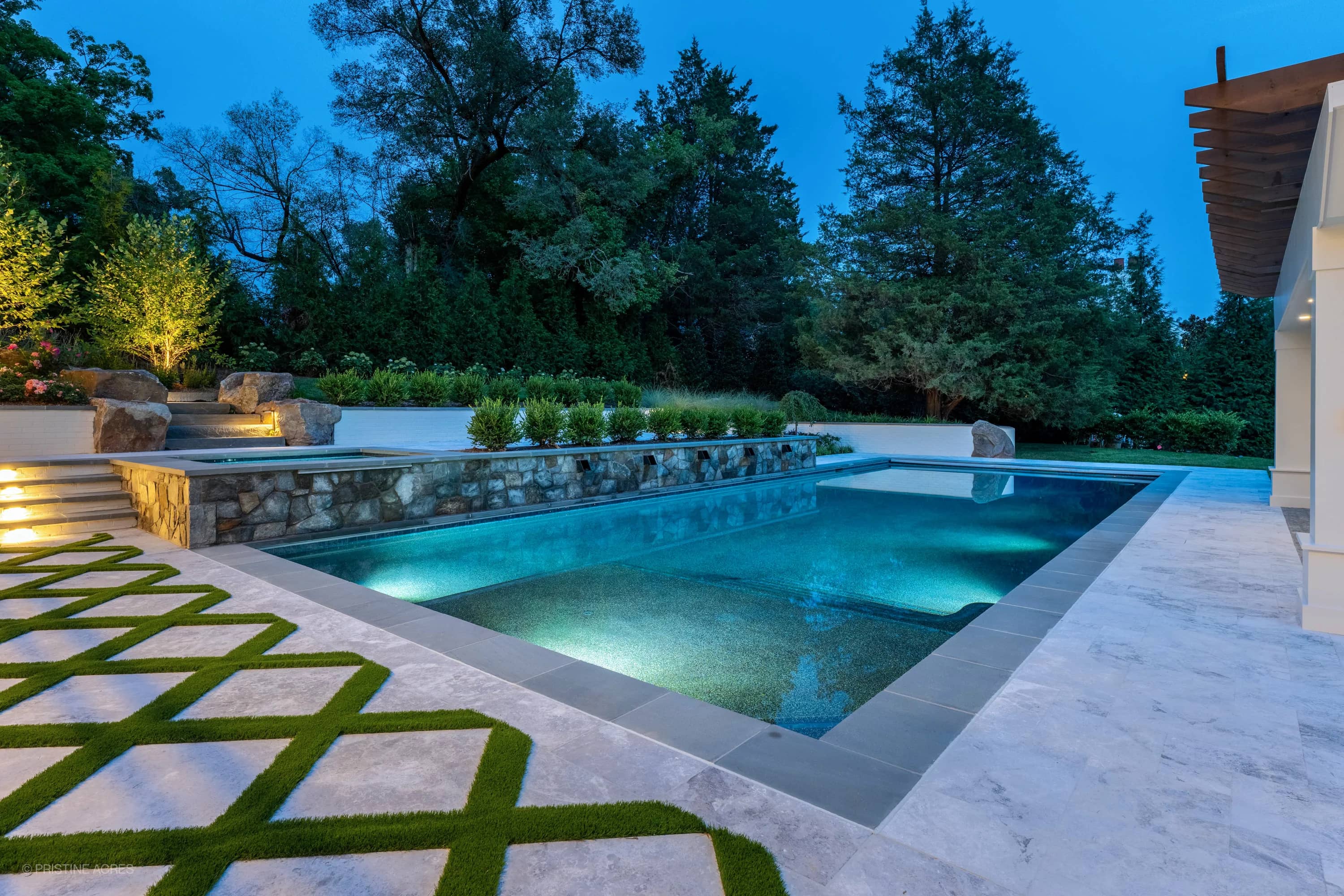 Luxurious pool deck in McLean, VA featuring Tundra Grey Marble pavers and Bluestone coping. The modern design includes a turquoise pool, stone accents, lush landscaping, and elegant outdoor lighting, creating a serene backyard oasis.