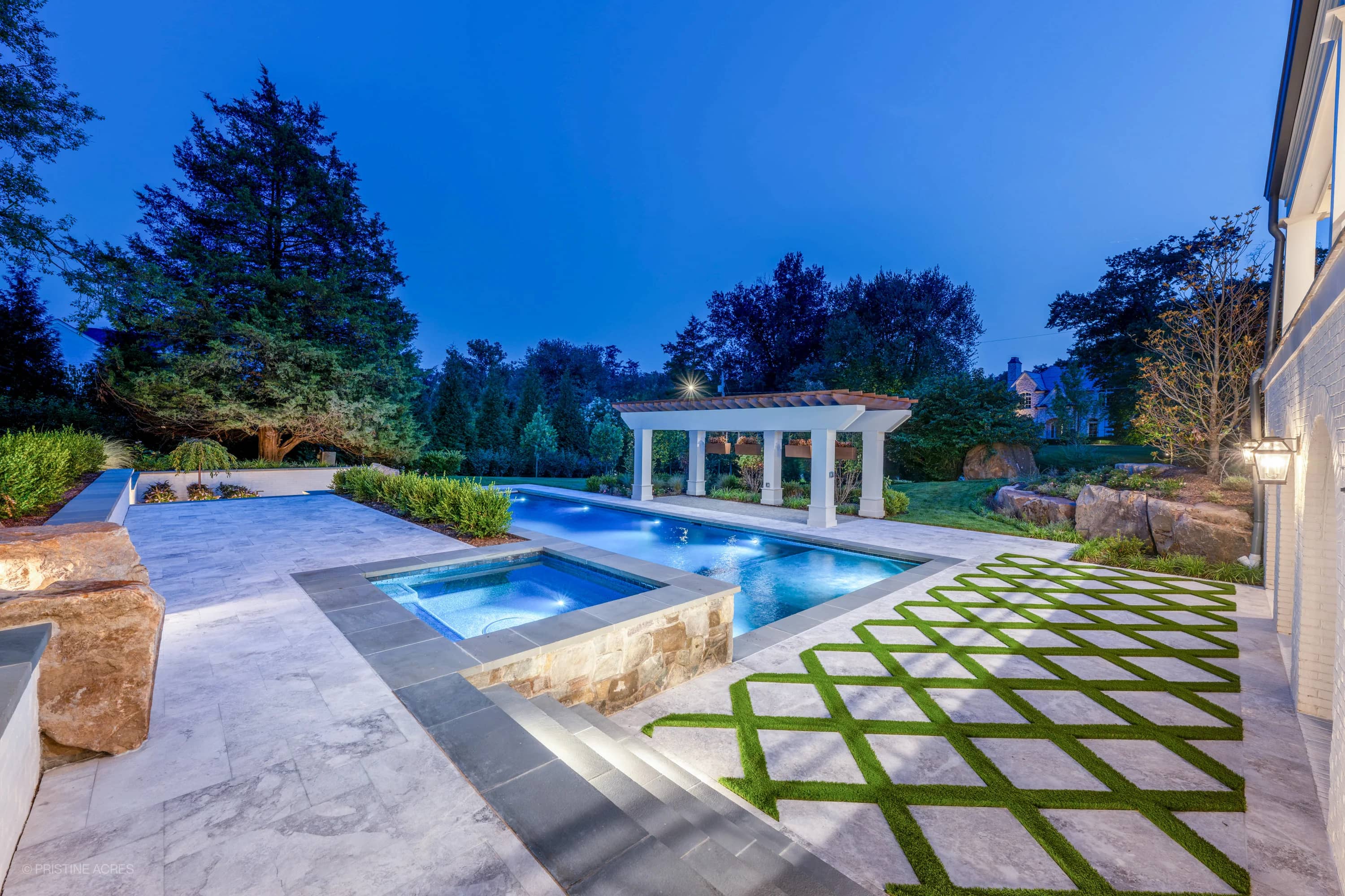 Luxurious pool deck featuring Tundra Grey Marble from STONELUX in McLean, VA. The bluestone coping elegantly complements the blue pool water. Surrounded by lush greenery, modern architecture, and ambient evening lighting, creating a serene outdoor retr...